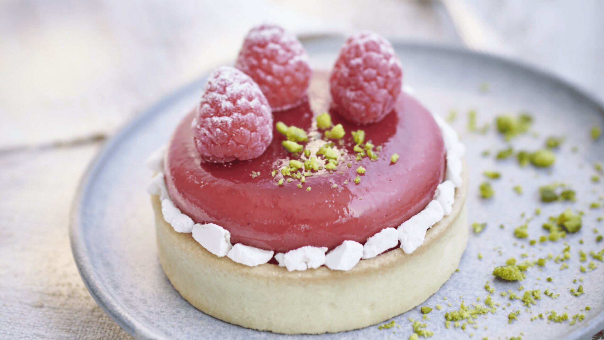 Eine Himbeertorte mit drei frischen Himbeeren, Puderzucker und zerstoßenen Pistazien liegt auf einem Teller, mit kleinen weißen Baiserstücken am Rand und ein paar Pistazienkrümeln auf dem Teller.