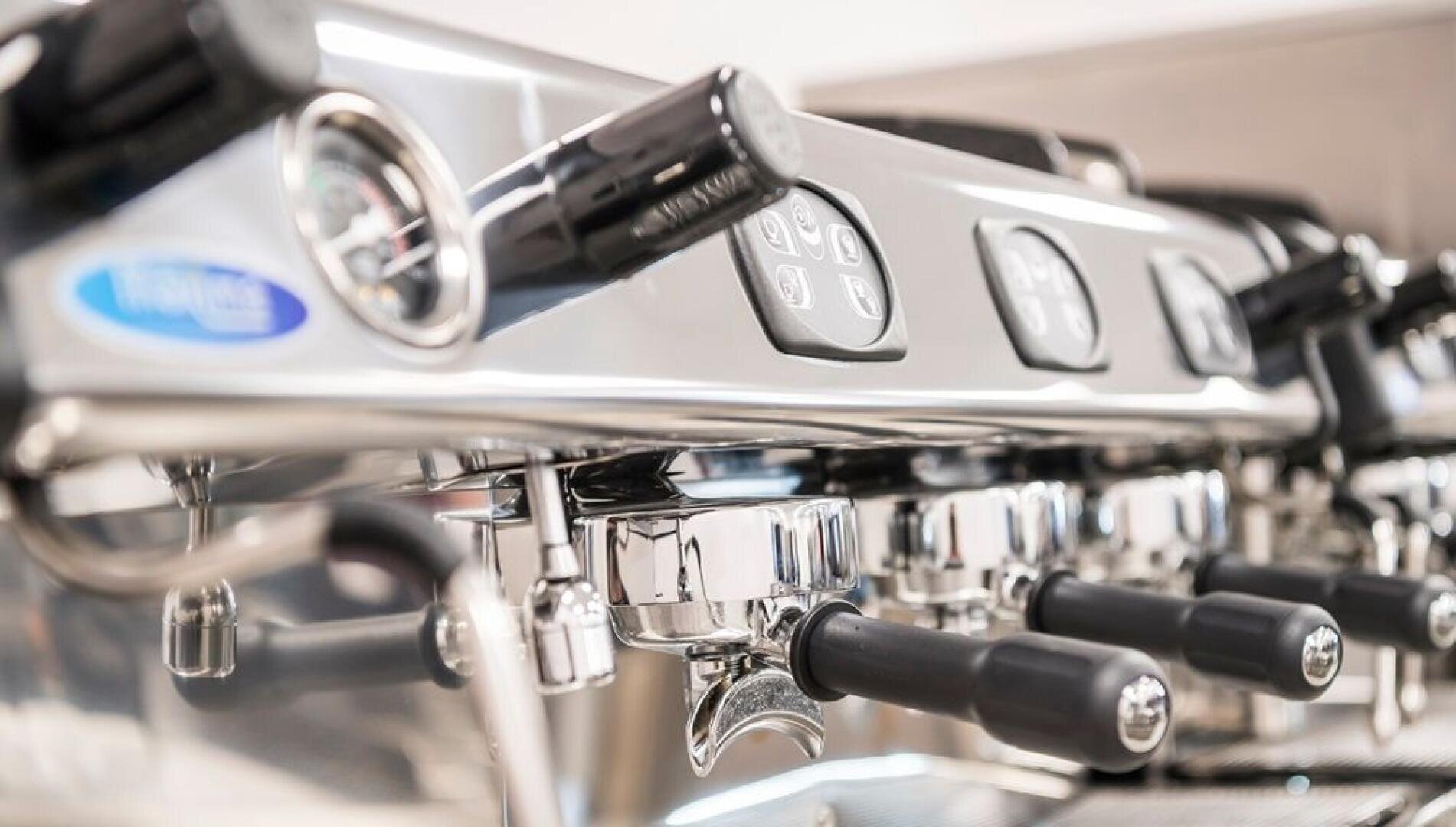Nahaufnahme einer handelsüblichen Espressomaschine mit glänzenden Chromteilen, mehreren Gruppenköpfen, Siebträgern und schwarzen Griffen, auf denen Bedientasten und ein Druckmesser zu sehen sind.