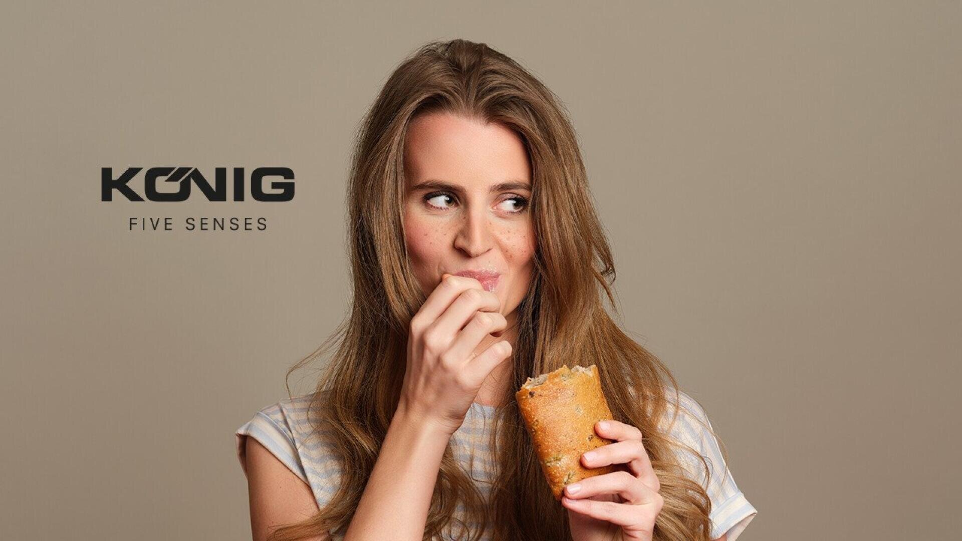 A woman with long, light brown hair is smiling and looking to the side while eating a wrap. The words König Five Senses are displayed to her left on a plain background.