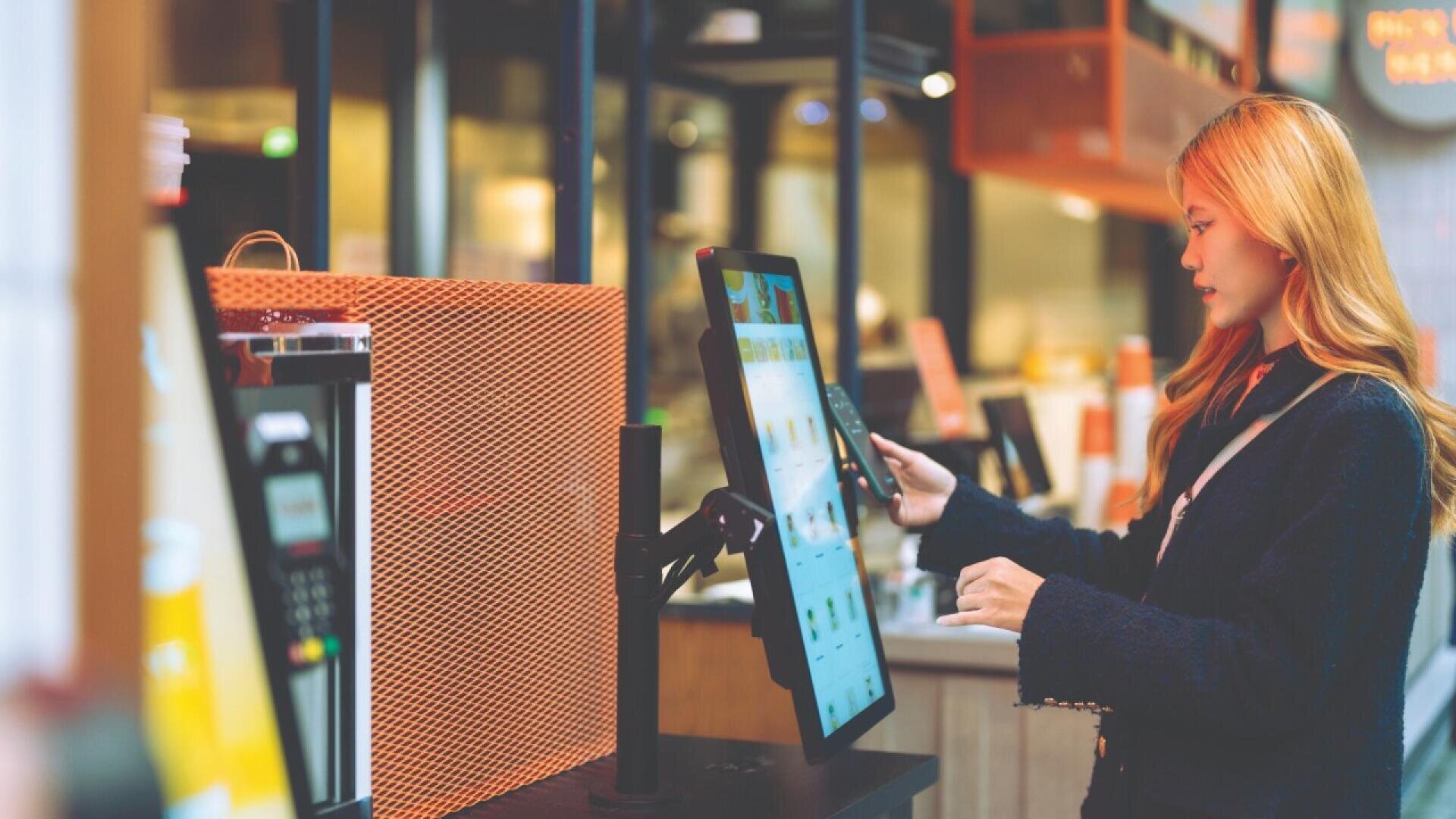 A woman uses her smartphone to make a payment at a touchscreen self-service kiosk in a modern, brightly lit store or café.