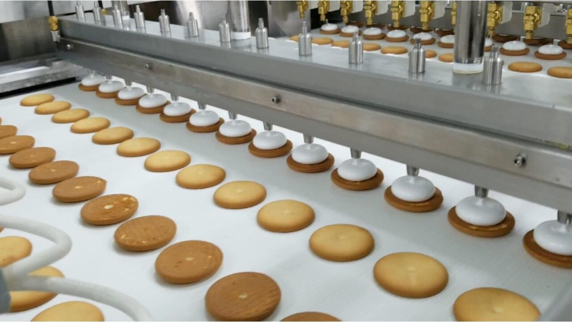 In einer Lebensmittelproduktionsanlage werden Kekse auf einem Förderband durch eine Reihe automatischer Düsen mit weißer Sahne bestrichen.