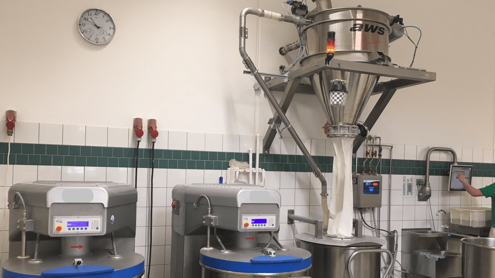 Industrieküche mit zwei großen Mischern, einer Uhr an der Wand, automatischen Geräten, die Zutaten von oben ausgeben, gefliesten Wänden und einer Person, die ein Touchscreen-Bedienfeld auf der rechten Seite bedient.