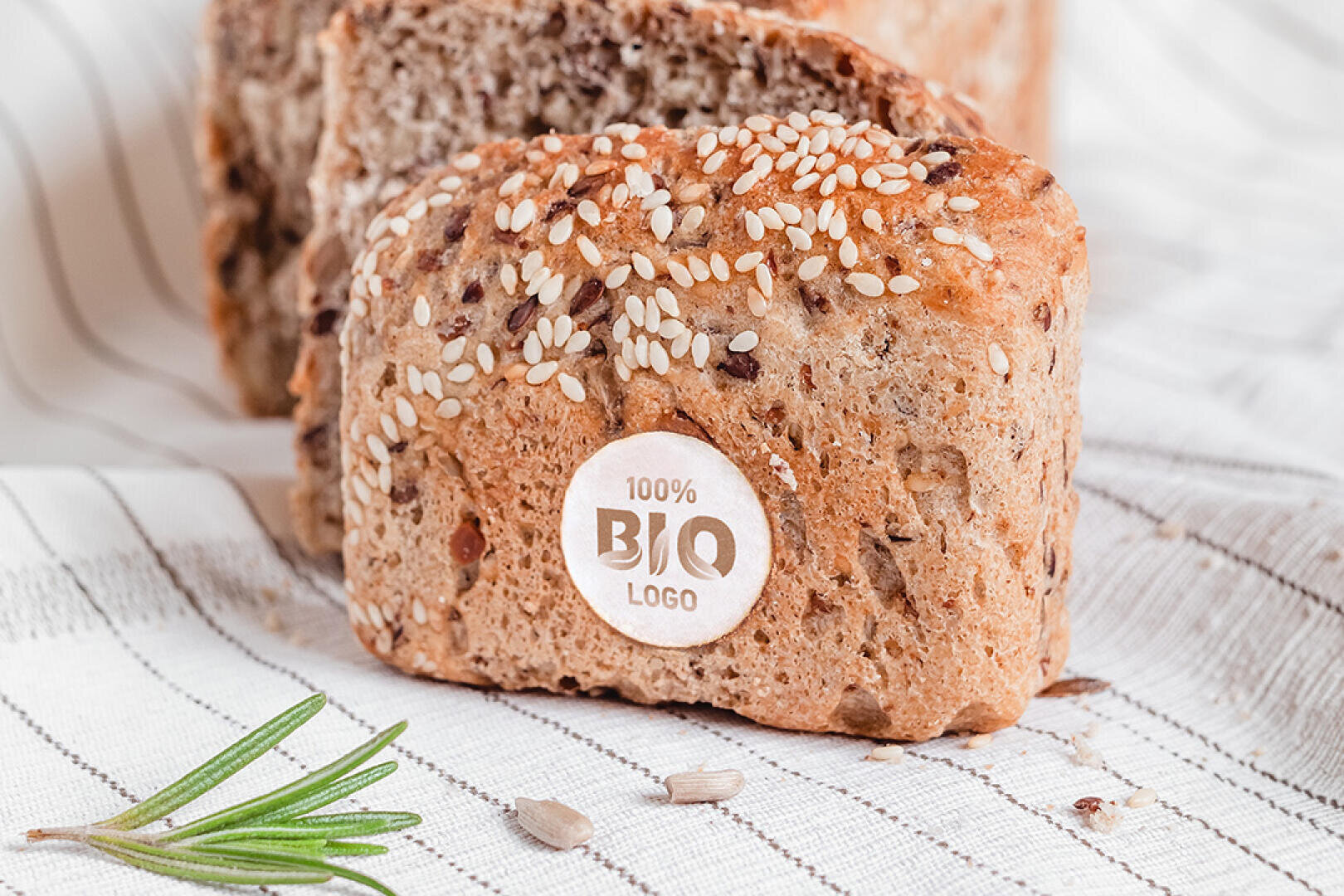 Nahaufnahme eines Laibes Vollkornbrot mit dem Etikett 100% BIO LOGO, der auf einem gestreiften Tuch liegt, mit einem Rosmarinzweig und verstreuten Samen in der Nähe.