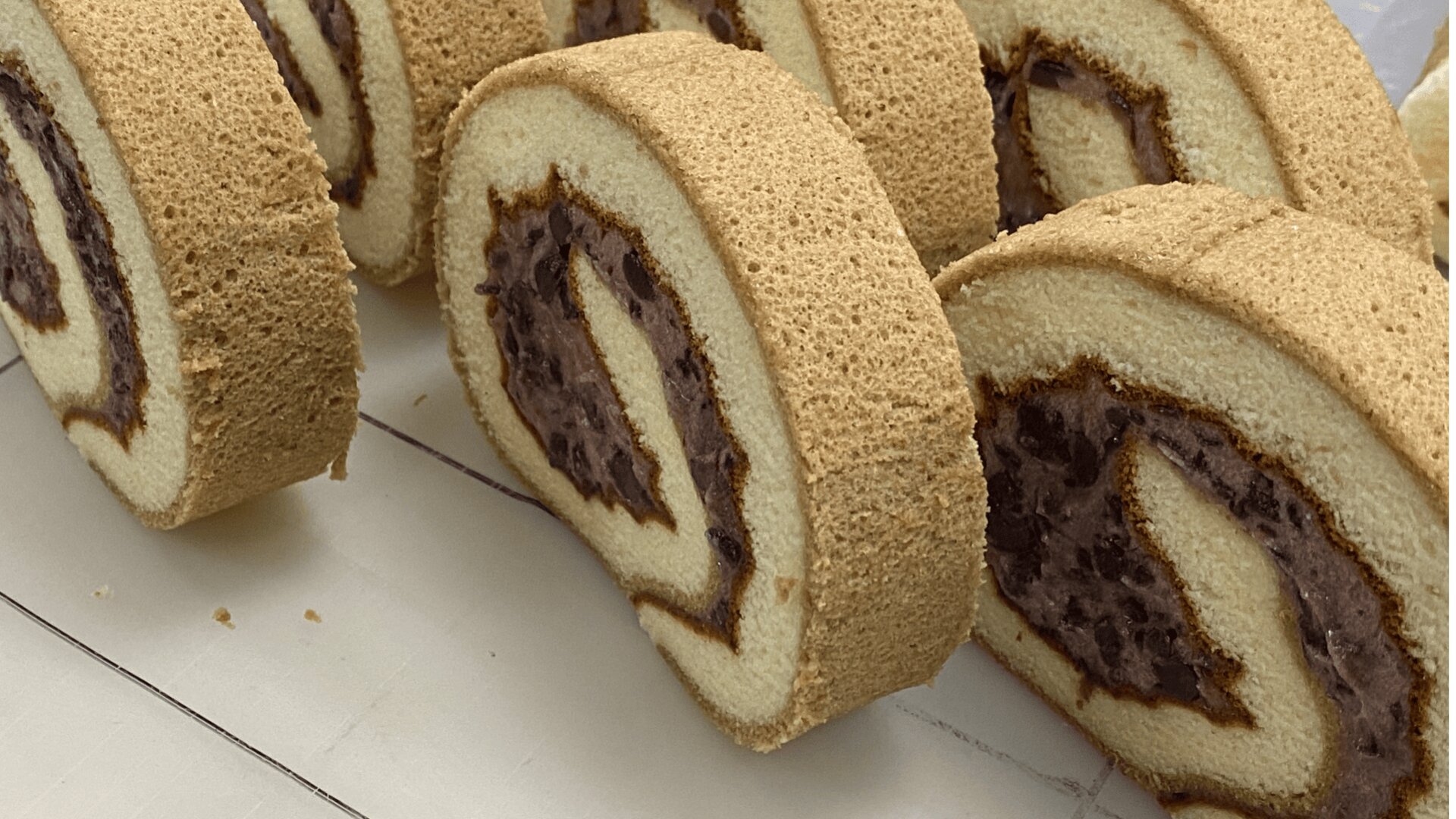 Sliced pieces of a rolled sponge cake with a dark red bean paste filling, neatly arranged on a light-colored surface. The cake is golden brown on the outside and has a visible swirl pattern.