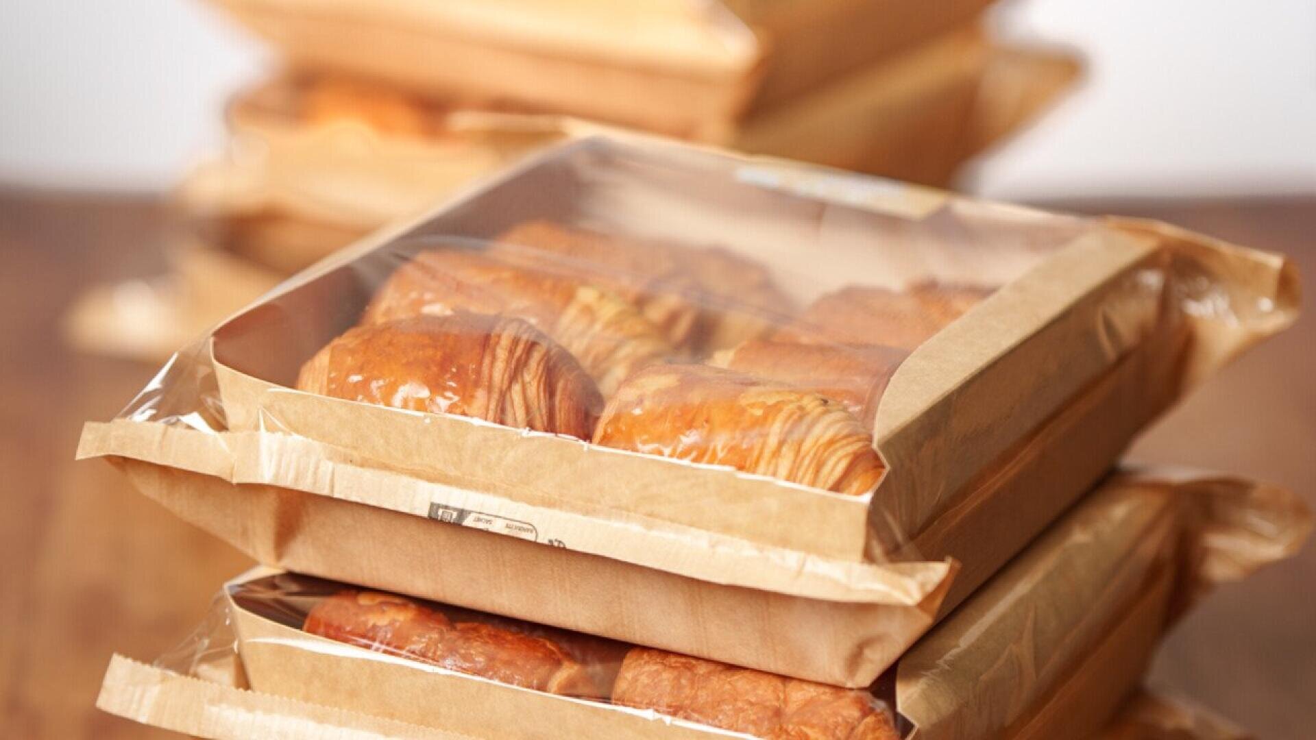 Drei Packungen Croissants in braunem Papier und durchsichtigen Plastikverpackungen sind auf einer hölzernen Oberfläche gestapelt, wobei weitere verpackte Backwaren im Hintergrund verschwimmen.