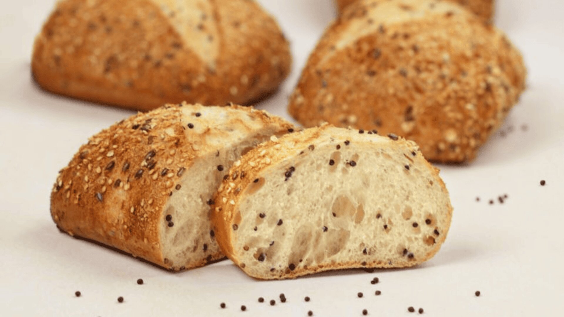 Four pieces of seeded bread are shown, with one piece sliced open to reveal its airy texture and seeds inside. Poppy and sesame seeds are scattered on the bread crust and nearby on a white surface.