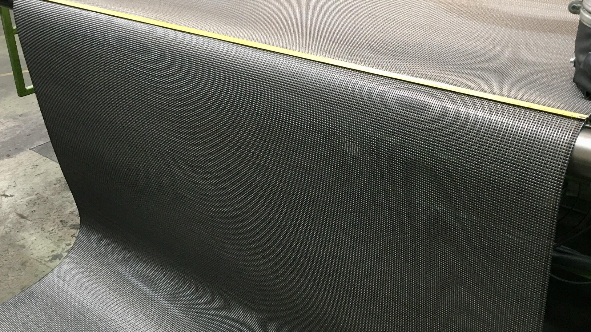 Eine große Rolle schwarzen Kohlefasergewebes wird in einer Werkstatt abgewickelt, wobei ein gelbes Maßband zur Orientierung über die Oberfläche gespannt ist.