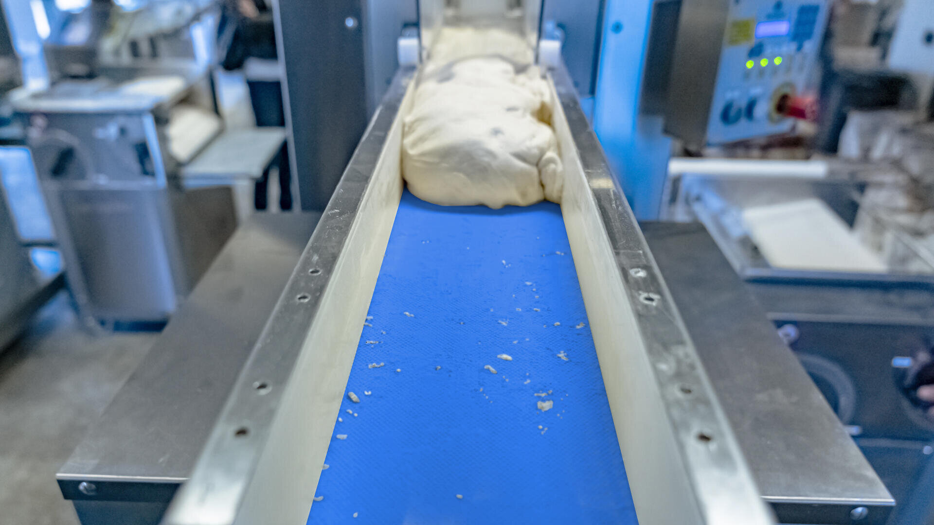 A lump of dough moves along a blue conveyor belt in a food processing facility, surrounded by industrial equipment and machinery.