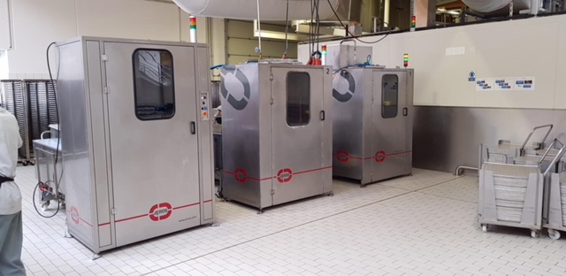 Drei große Industriemaschinen aus rostfreiem Stahl mit Glastüren stehen in einem sauberen, gefliesten Raum. In der Nähe befinden sich Stapel von Tabletts und Wagen, und an der Decke verlaufen Rohre.
