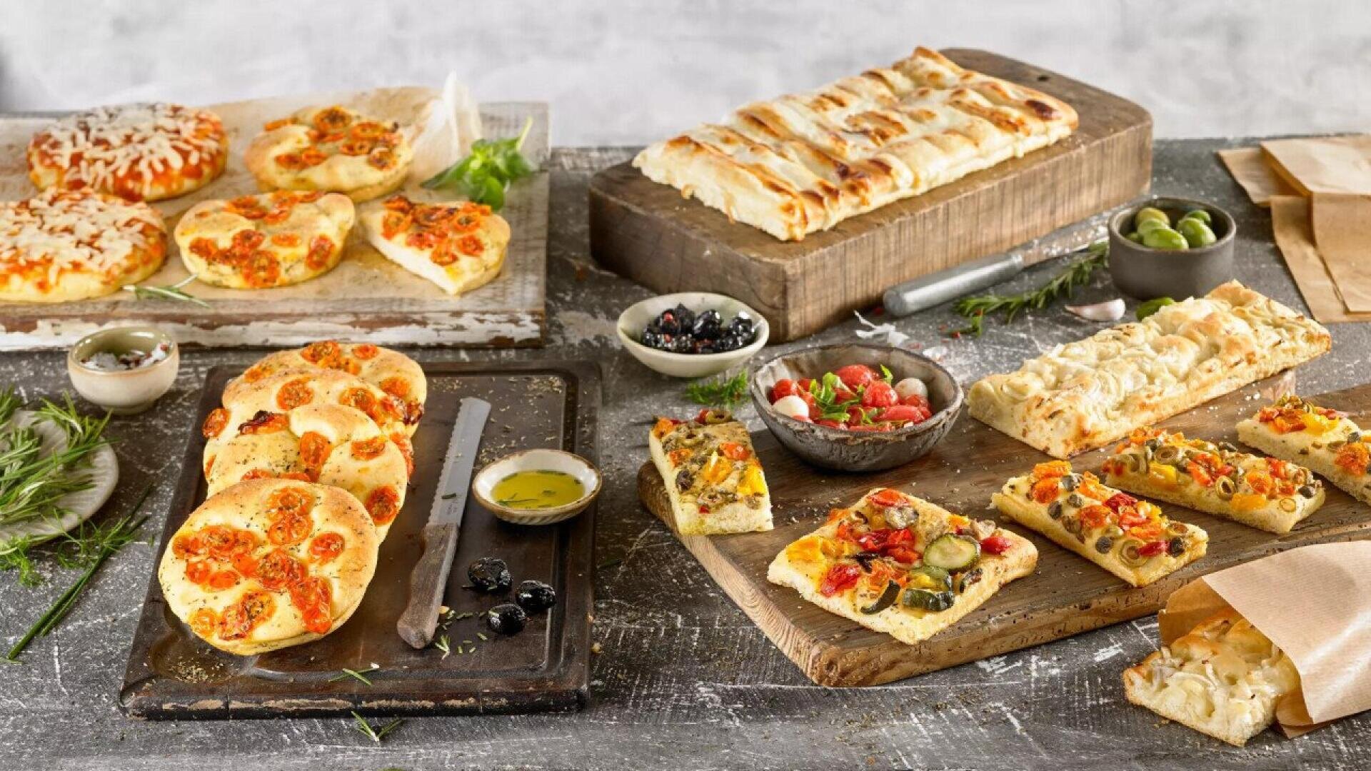 A rustic table displays various types of focaccia bread, some topped with tomatoes, olives, and vegetables, along with bowls of olives, a tomato salad, oil for dipping, and fresh herbs for garnish.