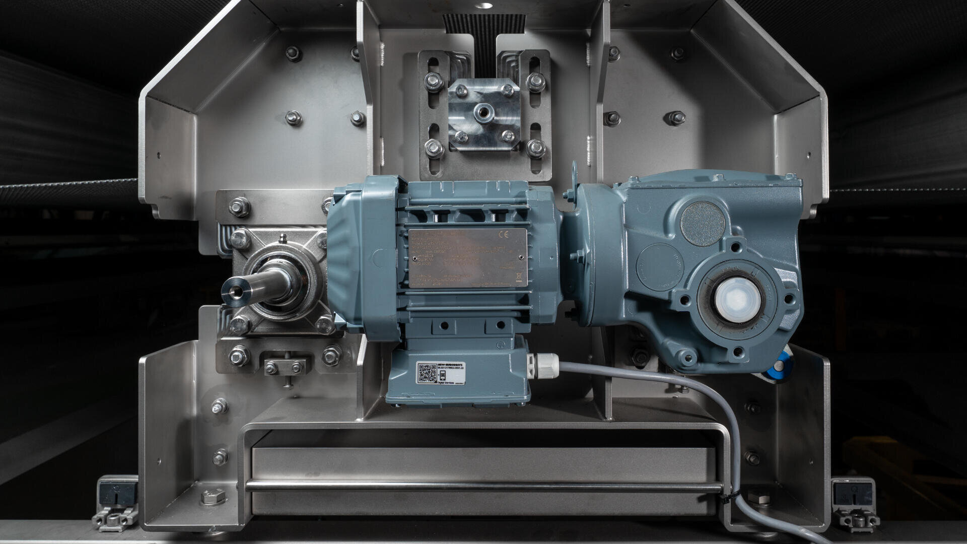 Nahaufnahme einer industriellen Elektromotor- und Getriebebaugruppe, die auf einem Metallrahmen montiert ist und verschiedene mechanische Komponenten und Kabel zeigt. Im Hintergrund sind weitere Metallteile und Bolzen zu sehen.