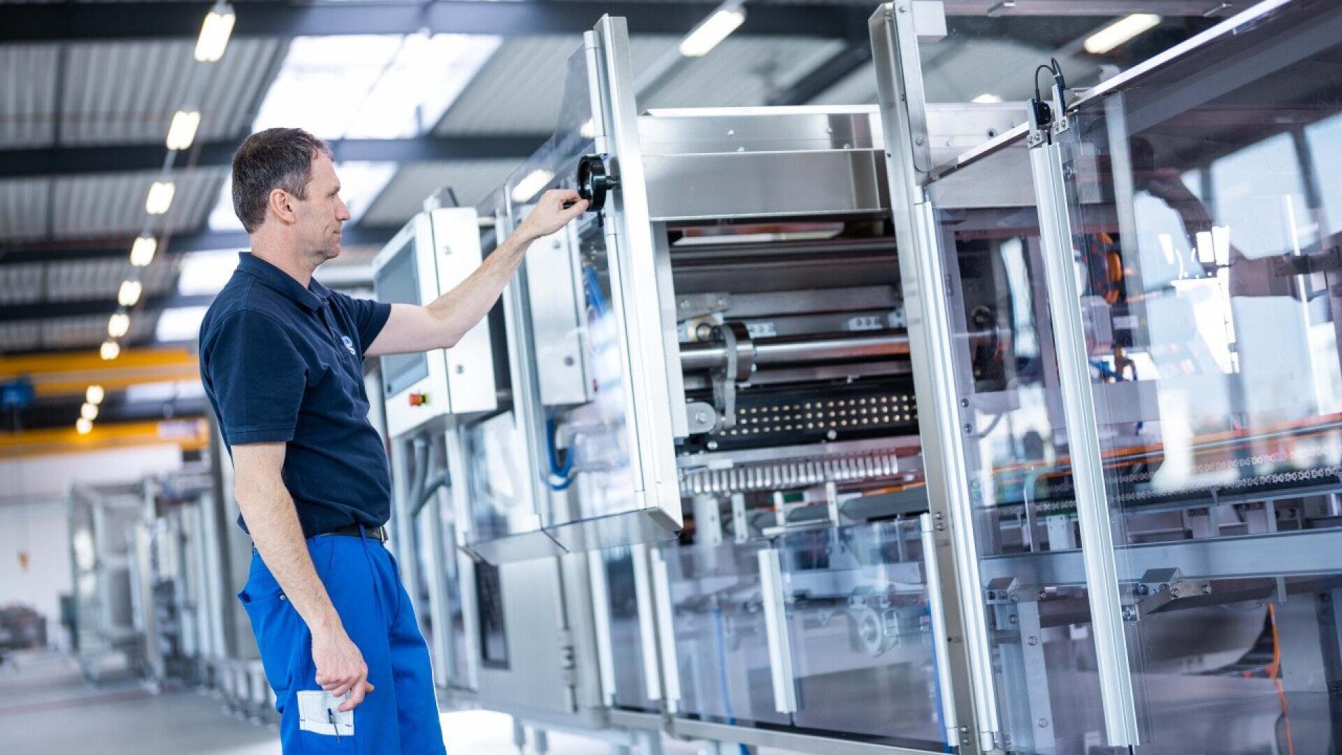 A man in blue work pants and a navy shirt operates a large industrial machine in a brightly lit factory with high ceilings and exposed beams.