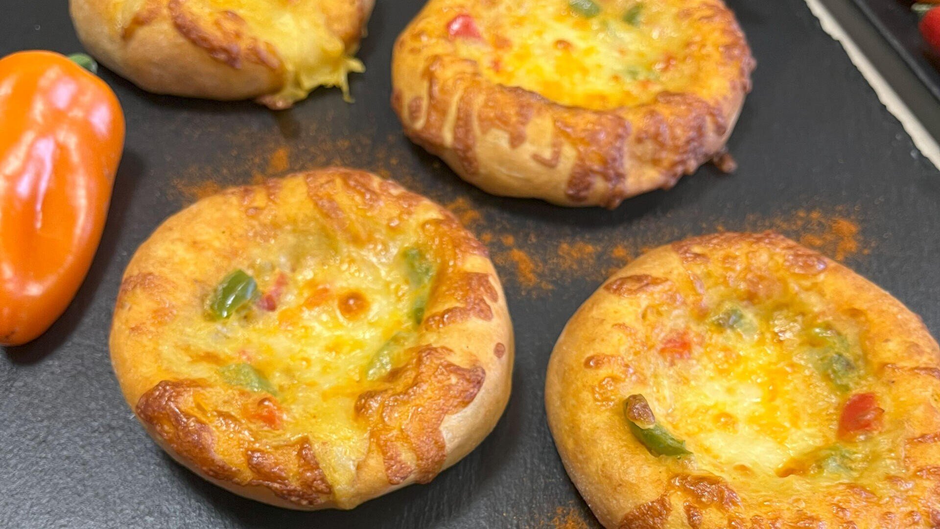 Vier kleine, runde Brötchen mit geschmolzenem Käse und gewürfelten roten und grünen Paprikaschoten liegen auf einer schwarzen Fläche neben einer orangefarbenen Paprika.
