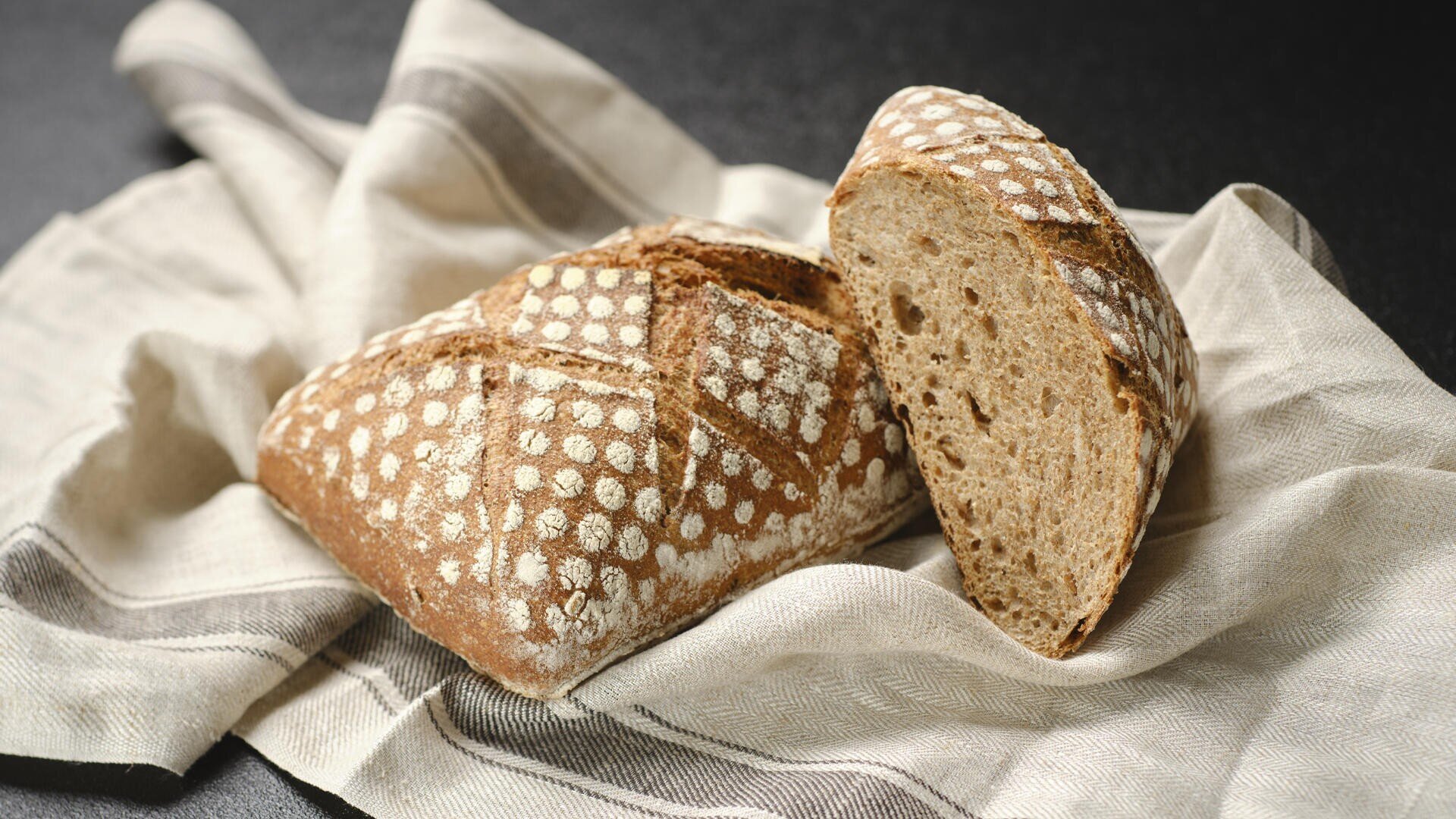 Ein rustikaler Brotlaib mit gemusterter Mehlkruste liegt auf einem beige-grau gestreiften Tuch, eine Scheibe ist abgeschnitten und gibt den Blick auf die weiche, luftige Krume im Inneren frei.