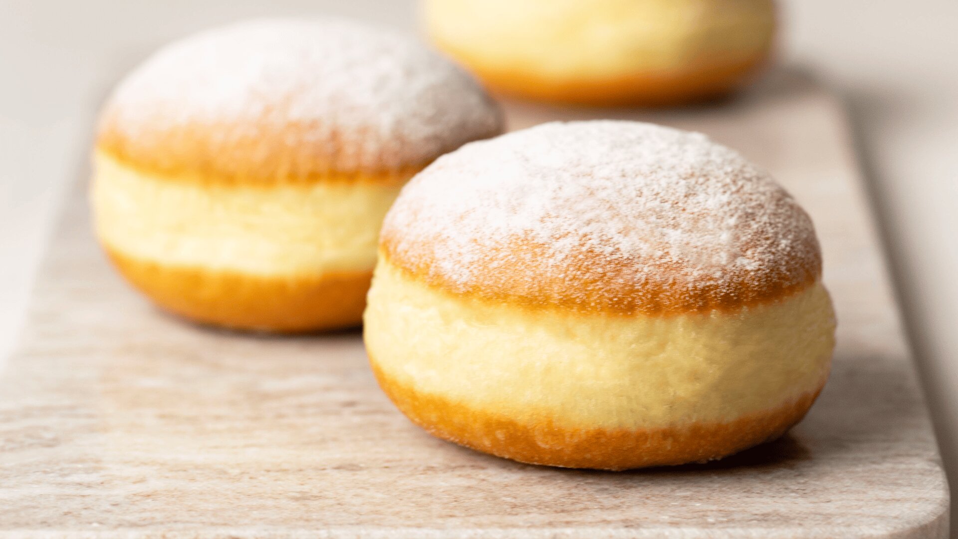 Drei runde, goldbraune, mit Puderzucker bestäubte Gelee-Krapfen werden auf einem hellen Holzbrett präsentiert. Der Schwerpunkt liegt auf dem vorderen Krapfen, der seine weiche Textur und sein fluffiges Aussehen zeigt.