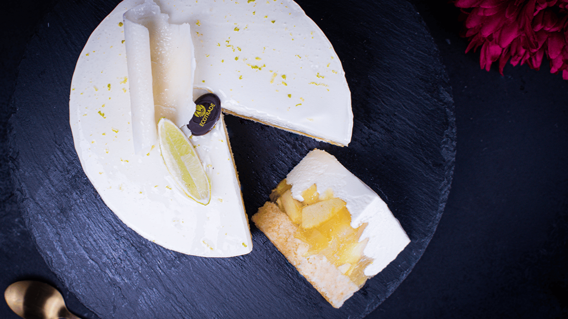 A round cheesecake garnished with a lime wedge and white chocolate curls sits on a dark slate plate. One slice is cut out, revealing a biscuit base and apple filling. A pink flower and gold spoon are nearby.