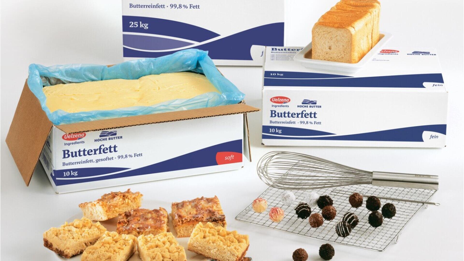 A large box of butterfat, labeled Butterfett, sits open with a slab of butter inside. Nearby are packaged loaves of bread, assorted pastries, chocolates, and a whisk on a cooling rack.