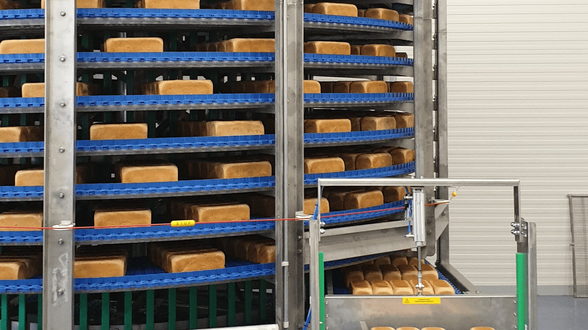 Metal racks filled with neatly arranged rectangular loaves of bread spiral upward in a commercial bakery, with a conveyor system moving bread in the foreground.