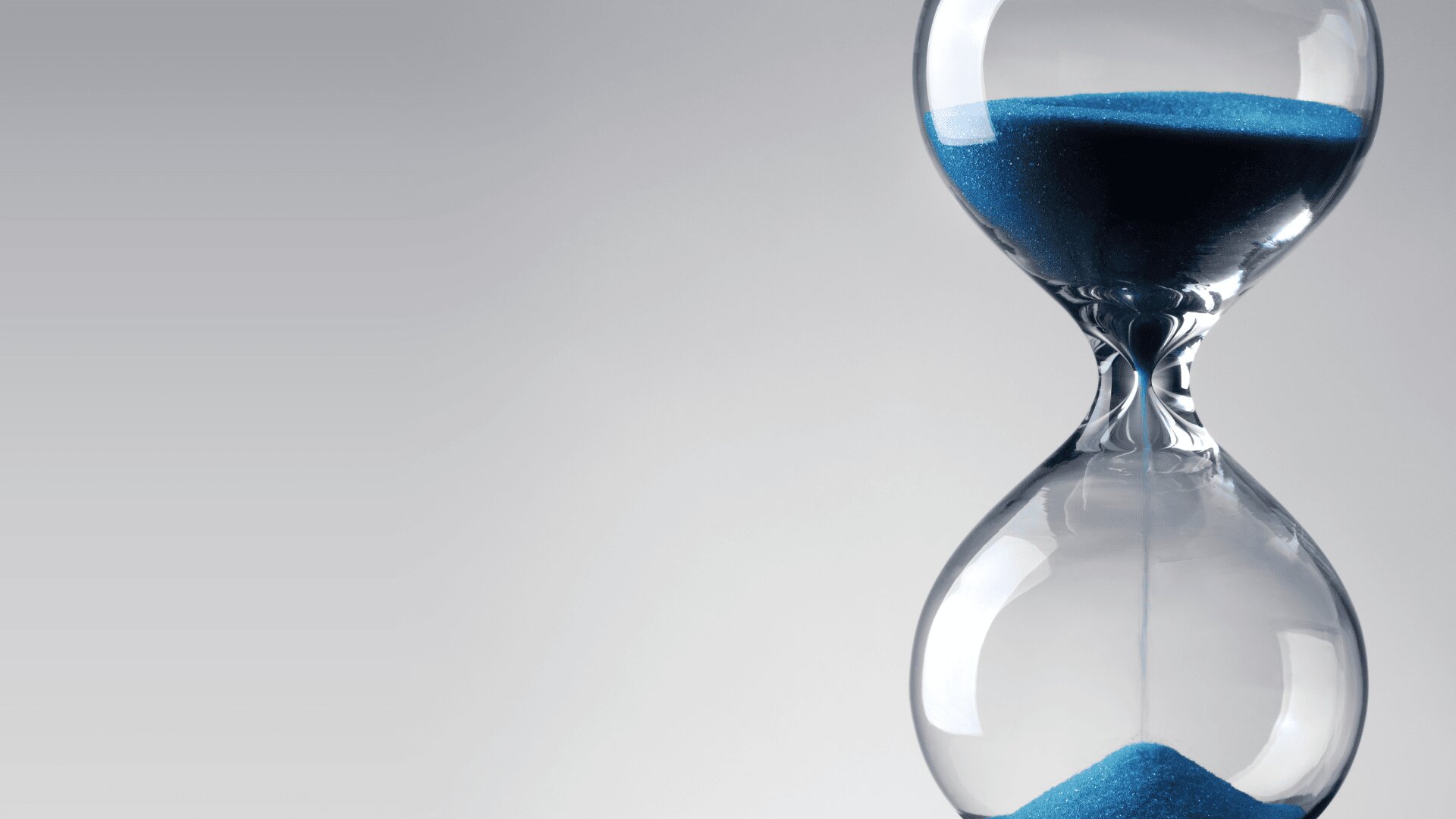 Eine gläserne Sanduhr mit blauem Sand, der von der oberen Kammer nach unten fließt, vor einem schlichten hellgrauen Hintergrund. Der Sand ist oben fast verschwunden.