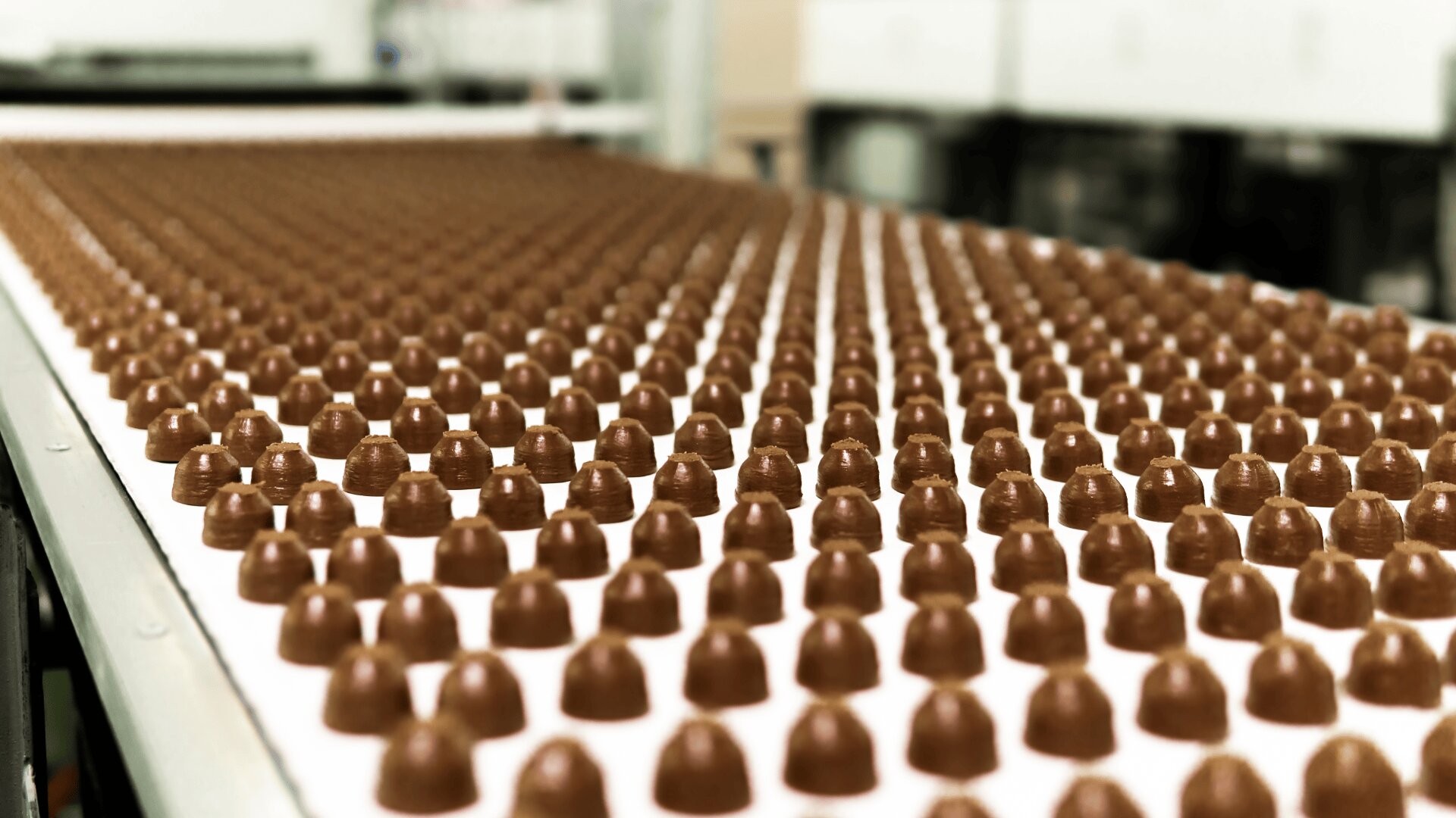 Rows of identical chocolate candies are arranged neatly on a conveyor belt in a factory setting, with a blurred background of industrial equipment.