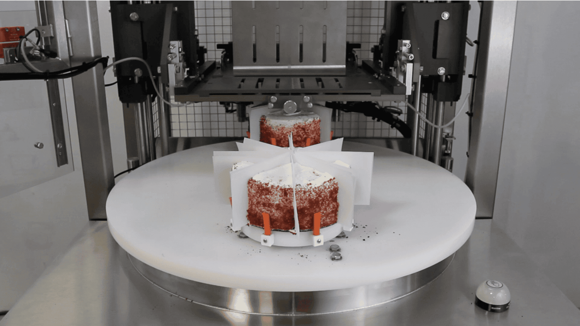 Eine Maschine, die einen runden, geschichteten Kuchen auf einer rotierenden Plattform mit vertikalen Messern und Führungen schneidet, um gleichmäßig große Stücke zu erzeugen. Auf der Oberfläche sind Krümel verstreut. Im Hintergrund sind Metall- und Gittertafeln zu sehen.