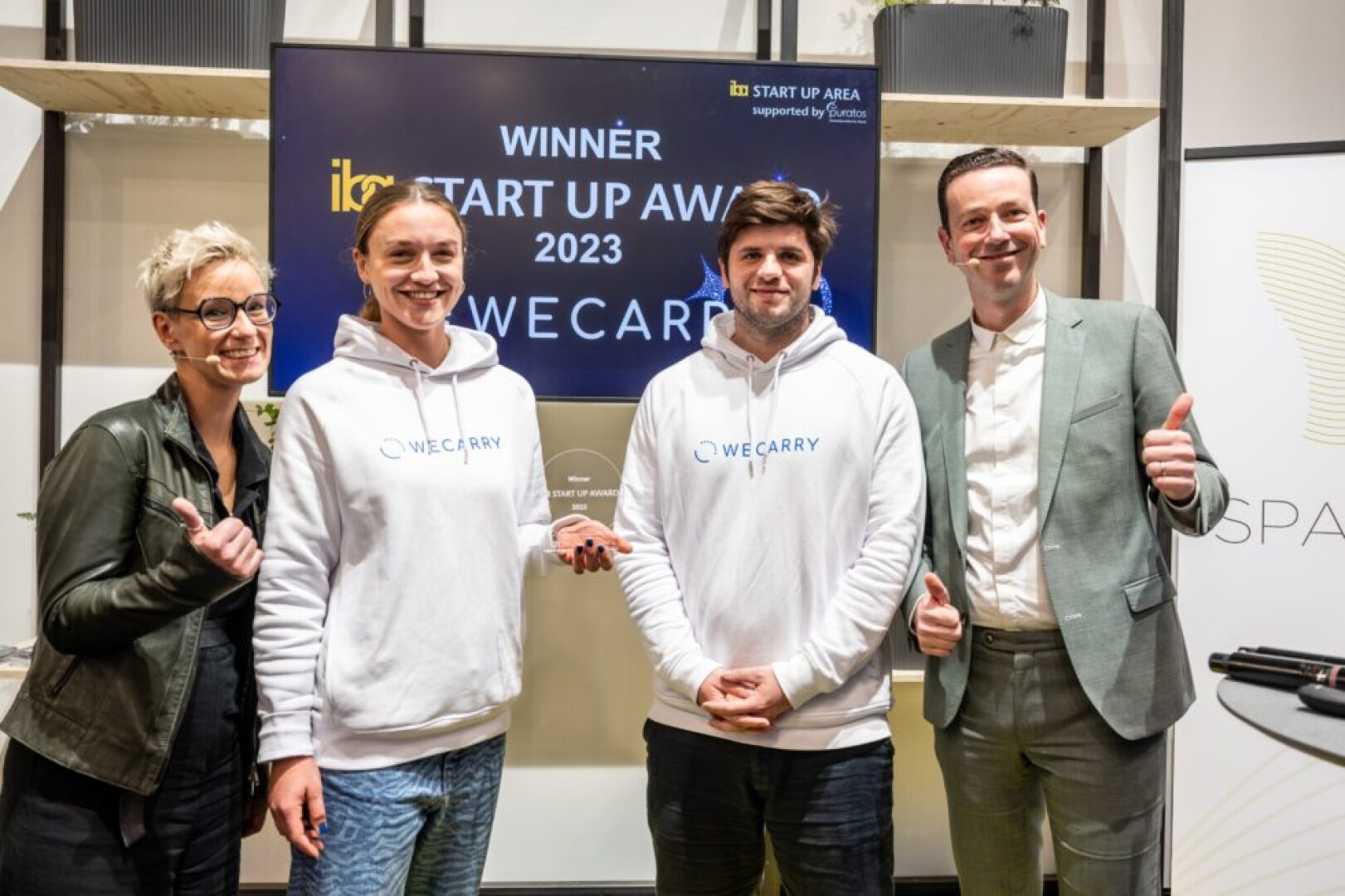 Vier Personen stehen lächelnd im Innenbereich; zwei in weißen WE CARRY-Hoodies halten Trophäen in den Händen. Auf einem Bildschirm hinter ihnen steht „WINNER START UP AWARD 2023 WE CARRY“. Die Gruppe posiert für ein Siegesfoto, einige strecken den Daumen hoch.