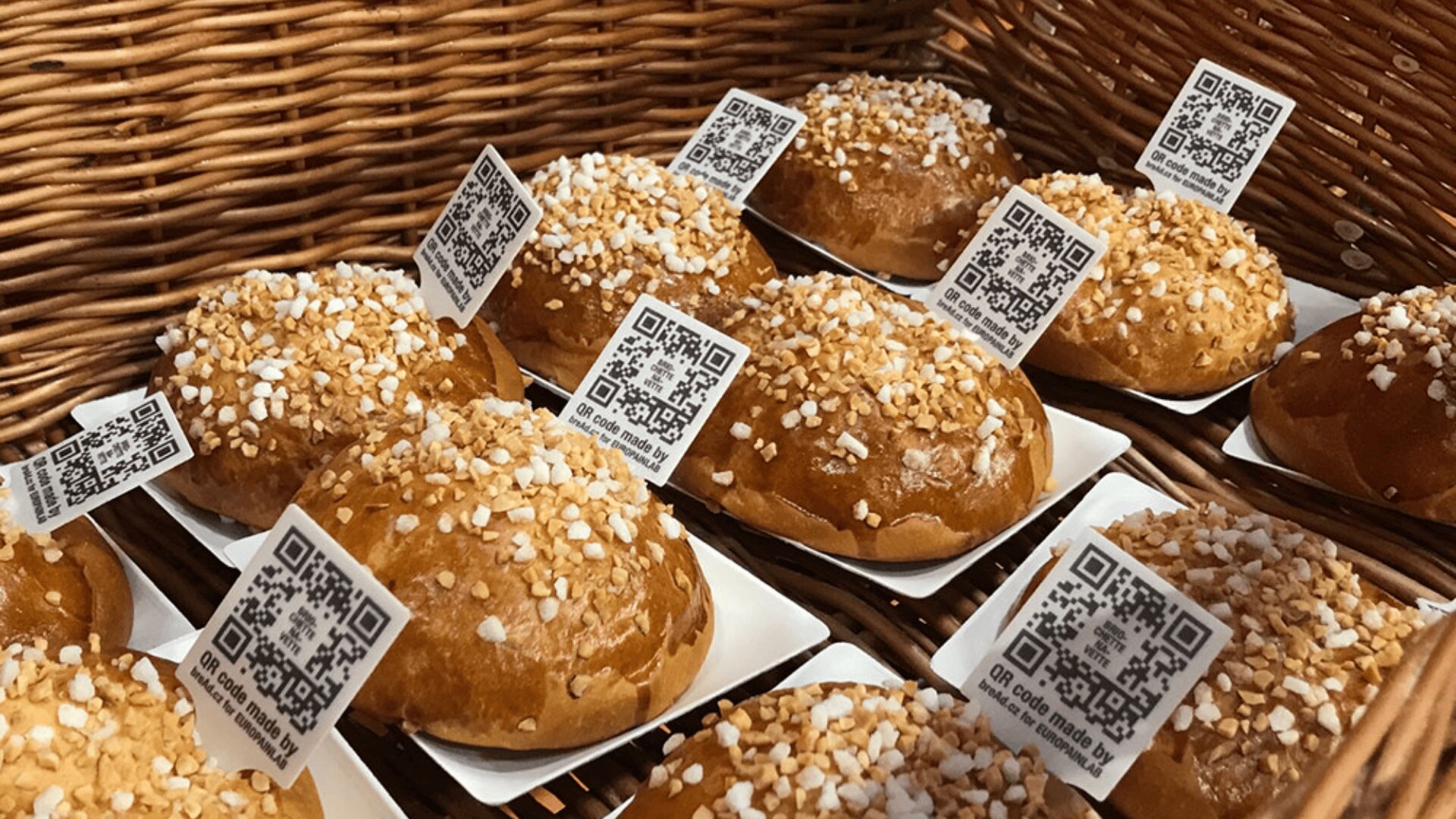 A basket filled with round pastries topped with pearl sugar, each sitting on a paper tray and labeled with a QR code card. The background is a woven wicker basket.