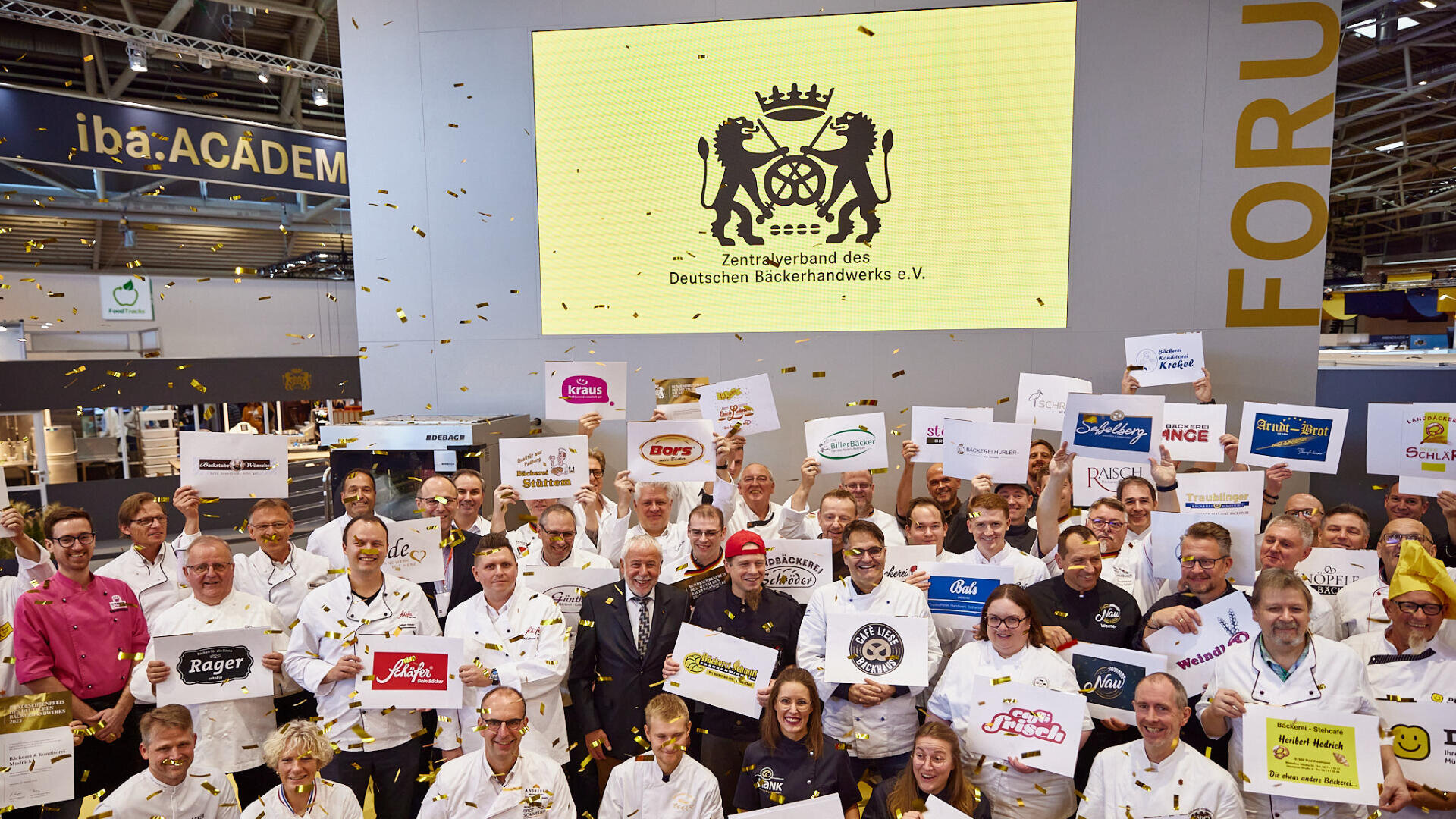 Eine große Gruppe von Menschen, viele in weißen Uniformen, steht in einem Raum und hält Schilder mit den Logos von Bäckereimarken. Von oben fällt goldenes Konfetti herab, und auf einem großen Bildschirm im Hintergrund ist das Logo des Deutschen Bäckerverbands zu sehen.