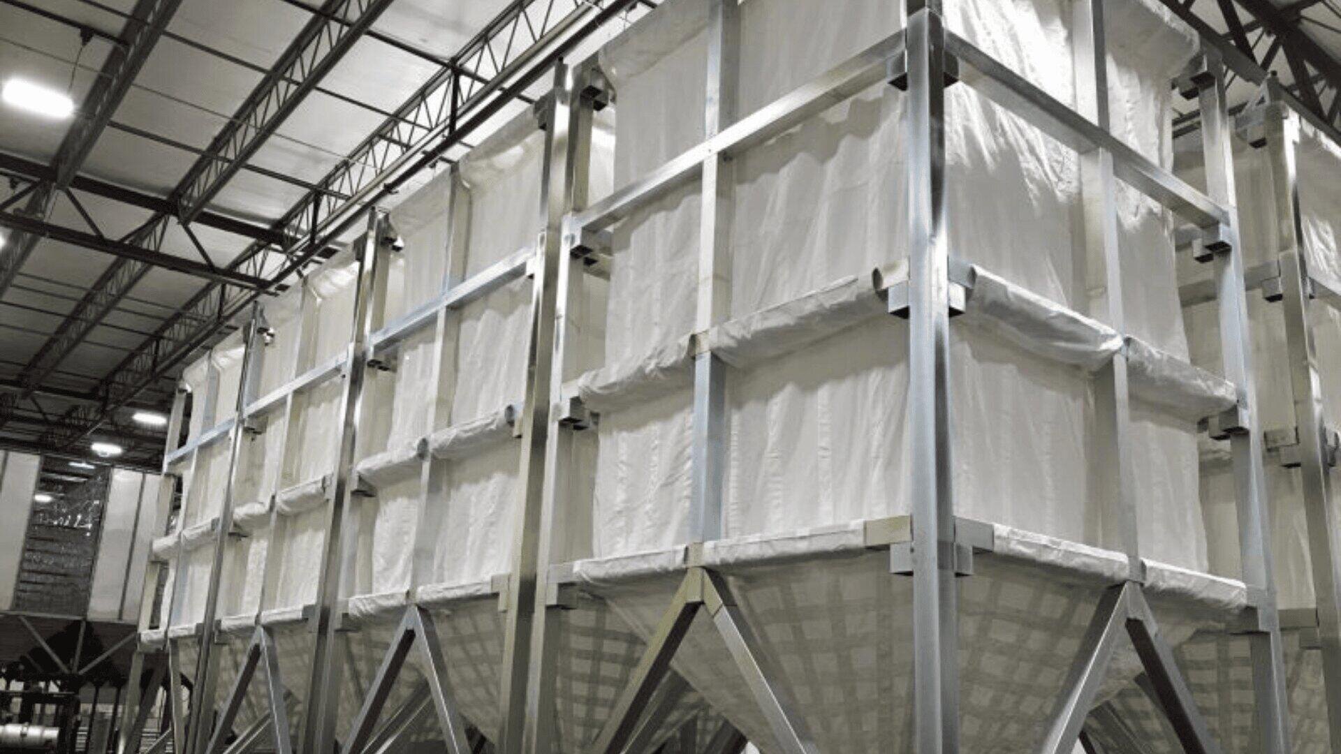 Large industrial fabric bulk storage bins with metal frames are arranged in rows inside a warehouse, under a high ceiling with visible steel beams and lighting.