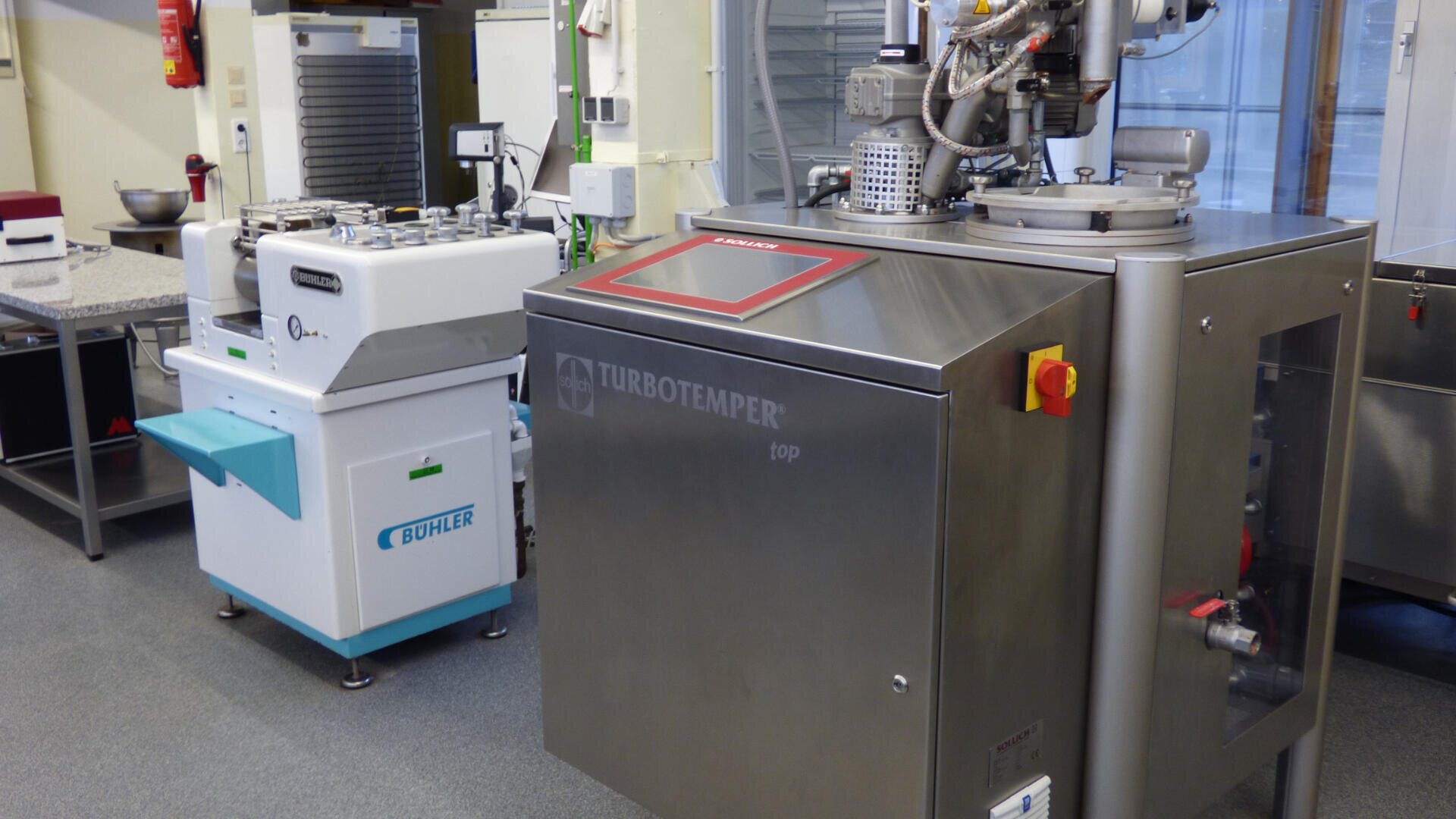Eine TURBOTEMPER-Maschine aus Edelstahl und eine weiße Bühler-Maschine stehen in einem hellen, modernen Labor mit grauen Böden und verschiedenen Geräten im Hintergrund.