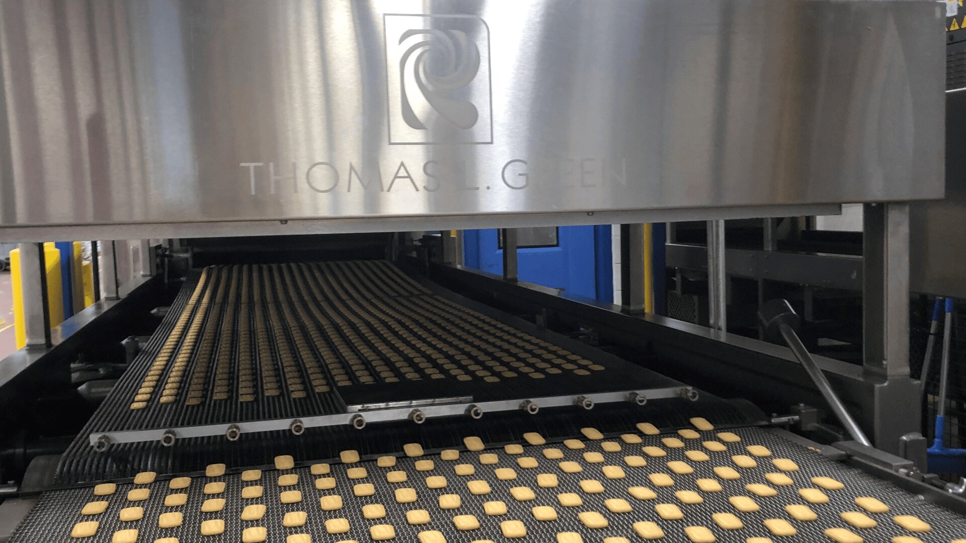 Rows of small square cookies move along a conveyor belt under a large stainless steel machine labeled THOMAS L. GREEN in a food production facility.