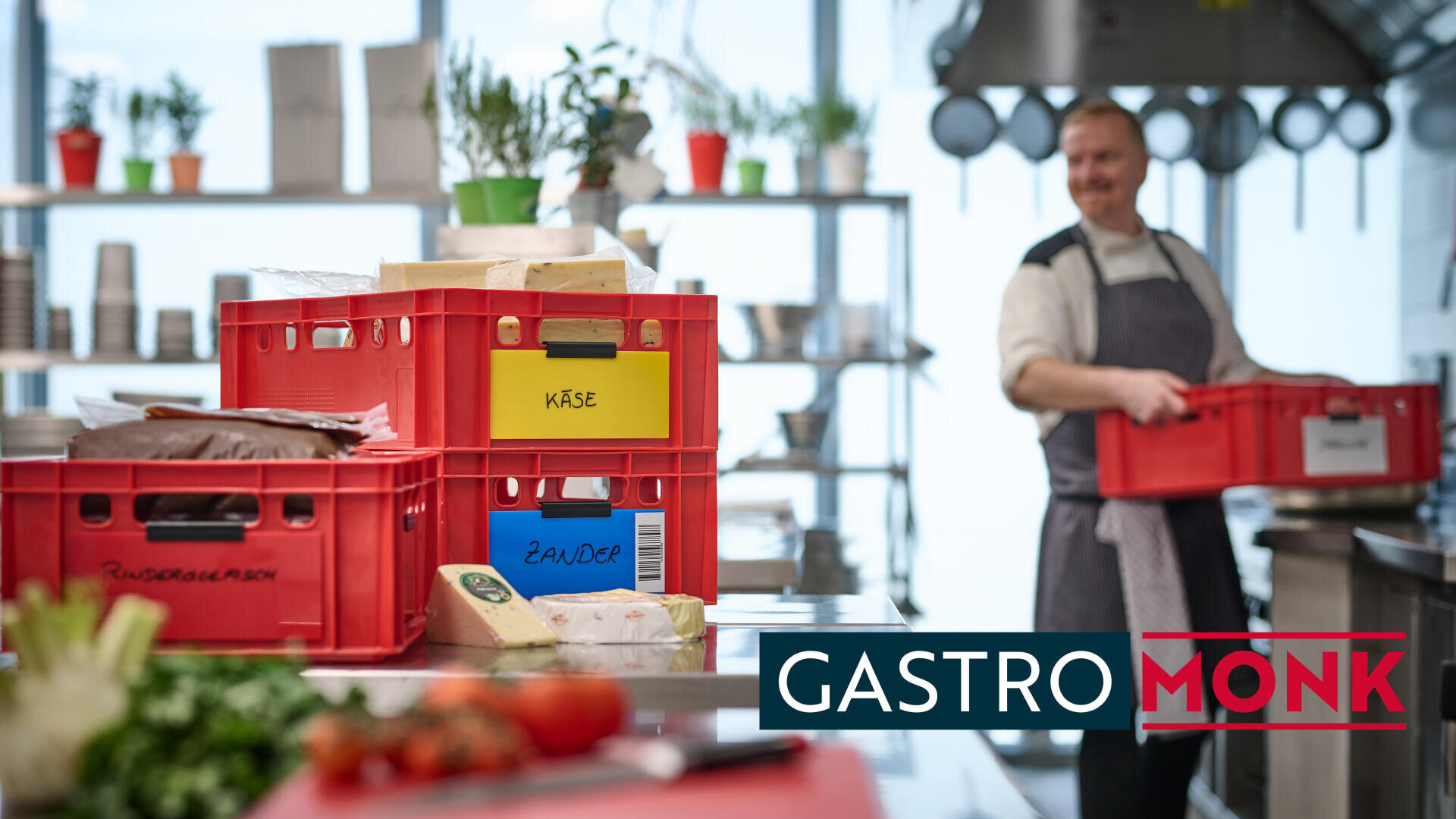 Ein Koch in einer Küche hält eine rote Kiste, umgeben von gestapelten Kisten mit der Aufschrift "Käse" und "Zander". Auf der Theke steht frisches Gemüse, und unten steht in fetter Schrift "GASTROMONK".