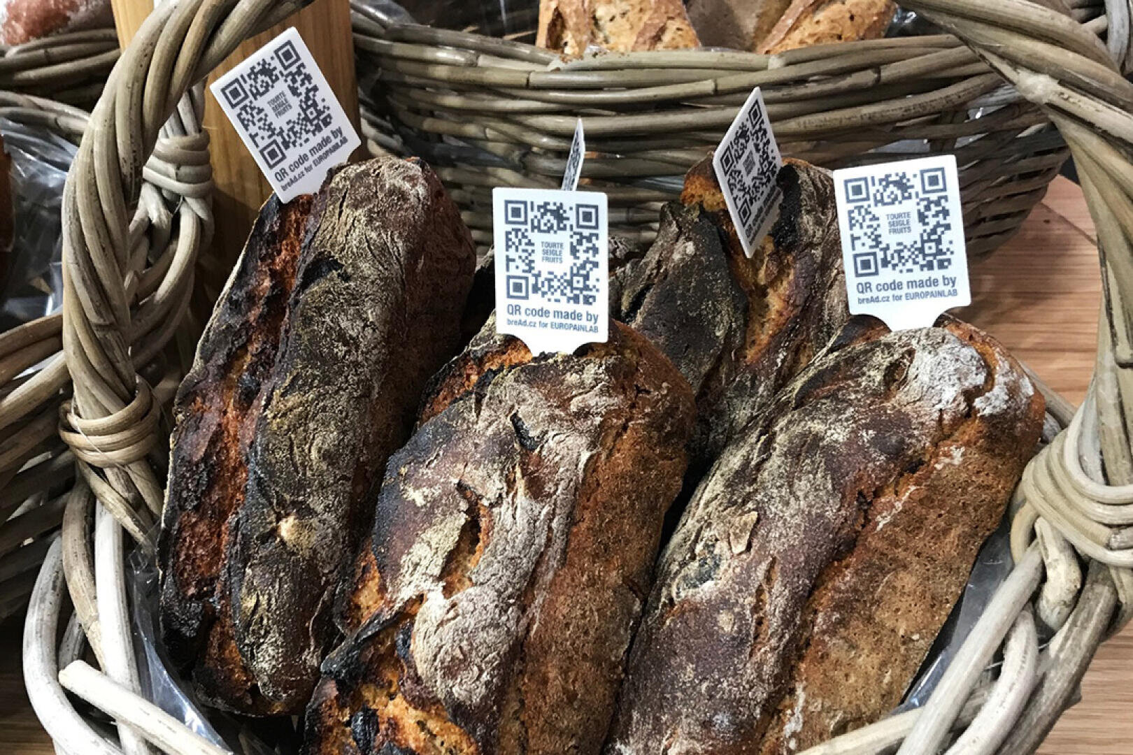 Ein Korb gefüllt mit rustikalen Broten, auf denen jeweils ein kleines Schild mit einem QR-Code für weitere Informationen angebracht ist. Im Hintergrund sind weitere Körbe mit Brot zu sehen.