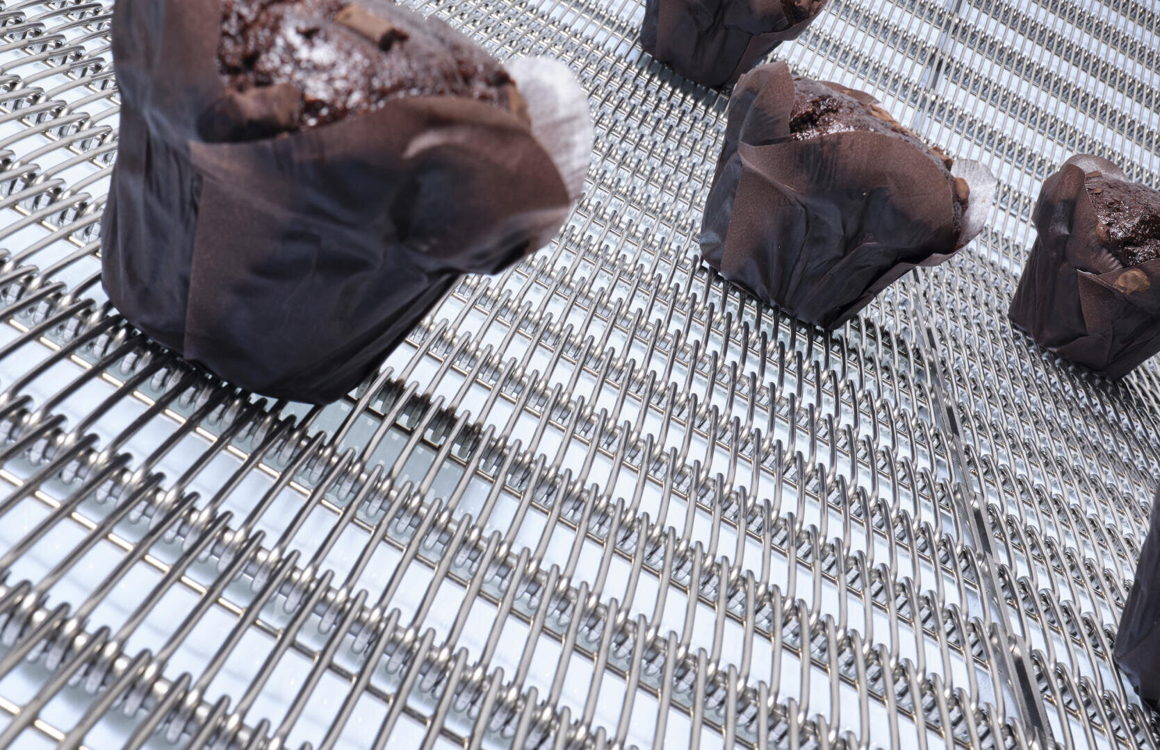 Schokoladenmuffins in braunen Verpackungen bewegen sich auf einem Metallförderband, wahrscheinlich in einer gewerblichen Bäckerei. Die Muffins liegen auf der Drahtgitteroberfläche in Abständen voneinander.