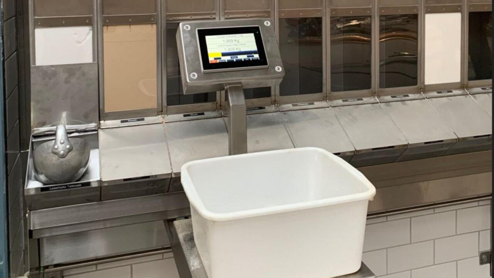 A digital scale with a display screen is mounted above a metal surface, holding a large empty white plastic bin. To the left, there is a metal figurine of a chicken near a tiled wall.
