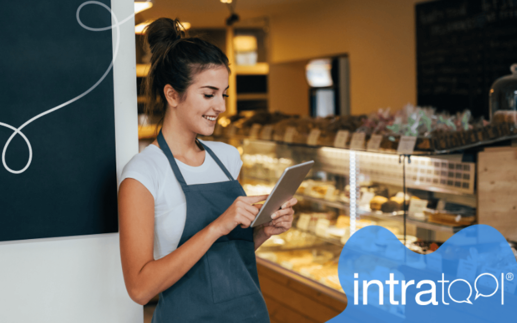 Eine Frau mit Schürze steht lächelnd in einer Bäckerei und bedient ein Tablet. Hinter ihr sind in Vitrinen Gebäckstücke ausgestellt. Unten rechts ist in einer blauen Grafik das Wort „intrataQI“ zu sehen.