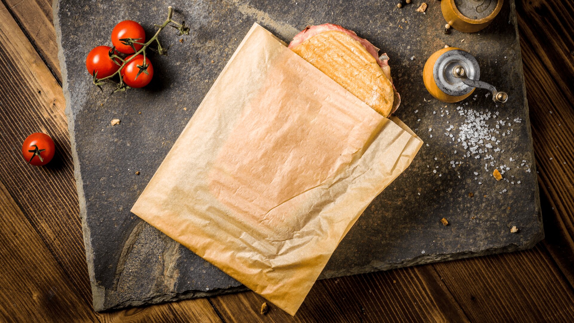 A sandwich partially wrapped in brown paper sits on a slate board, surrounded by cherry tomatoes on the vine, a salt shaker, some coarse salt, and a pepper mill on a wooden table.
