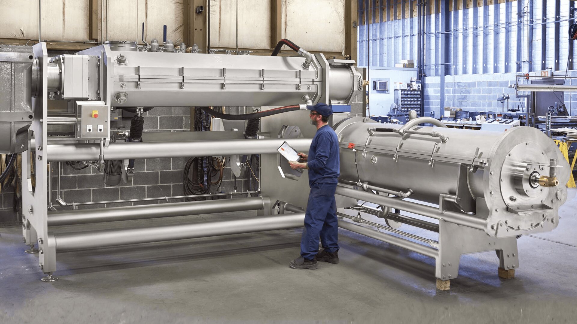 A worker in blue overalls and a cap inspects a large industrial stainless steel machine in a spacious factory setting, holding a tablet and standing on a concrete floor.