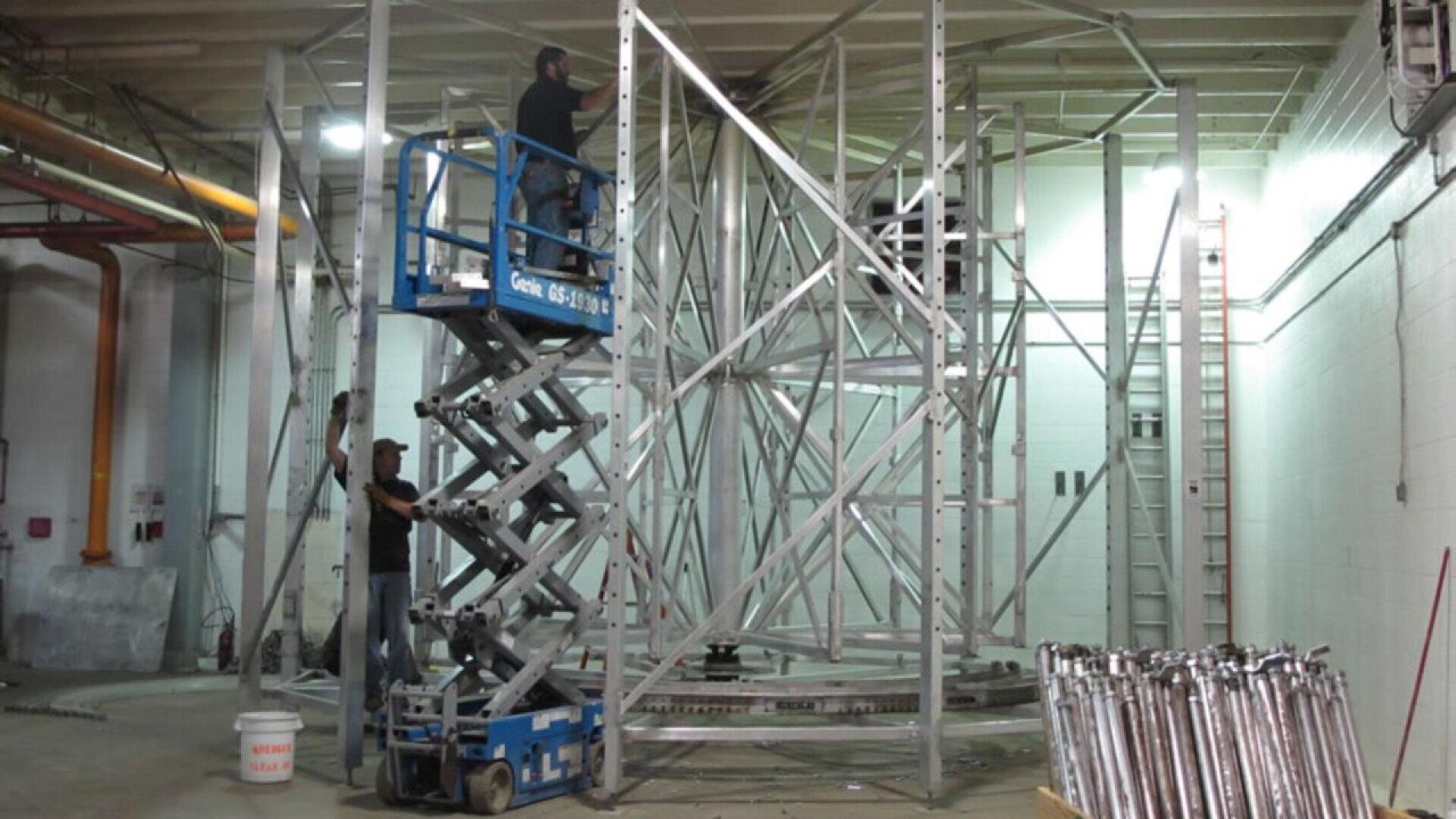 Zwei Arbeiter montieren in einer Halle eine große Metallstruktur; einer steht auf einer blauen Scherenhebebühne, der andere auf dem Boden. In dem Industrieraum sind Metallträger, Werkzeuge und eine Reihe von Rohren zu sehen.