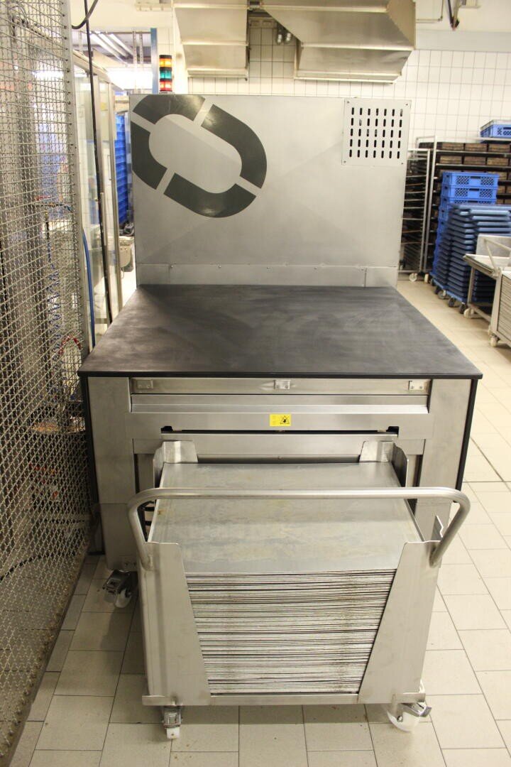 Eine Großküche mit einem Backofen oder einem Warmhaltegerät aus Edelstahl, einem ausgezogenen Tablettständer im Vordergrund und gestapelten Tabletts. Im Hintergrund sind blaue Regale und gekachelte Wände zu sehen.