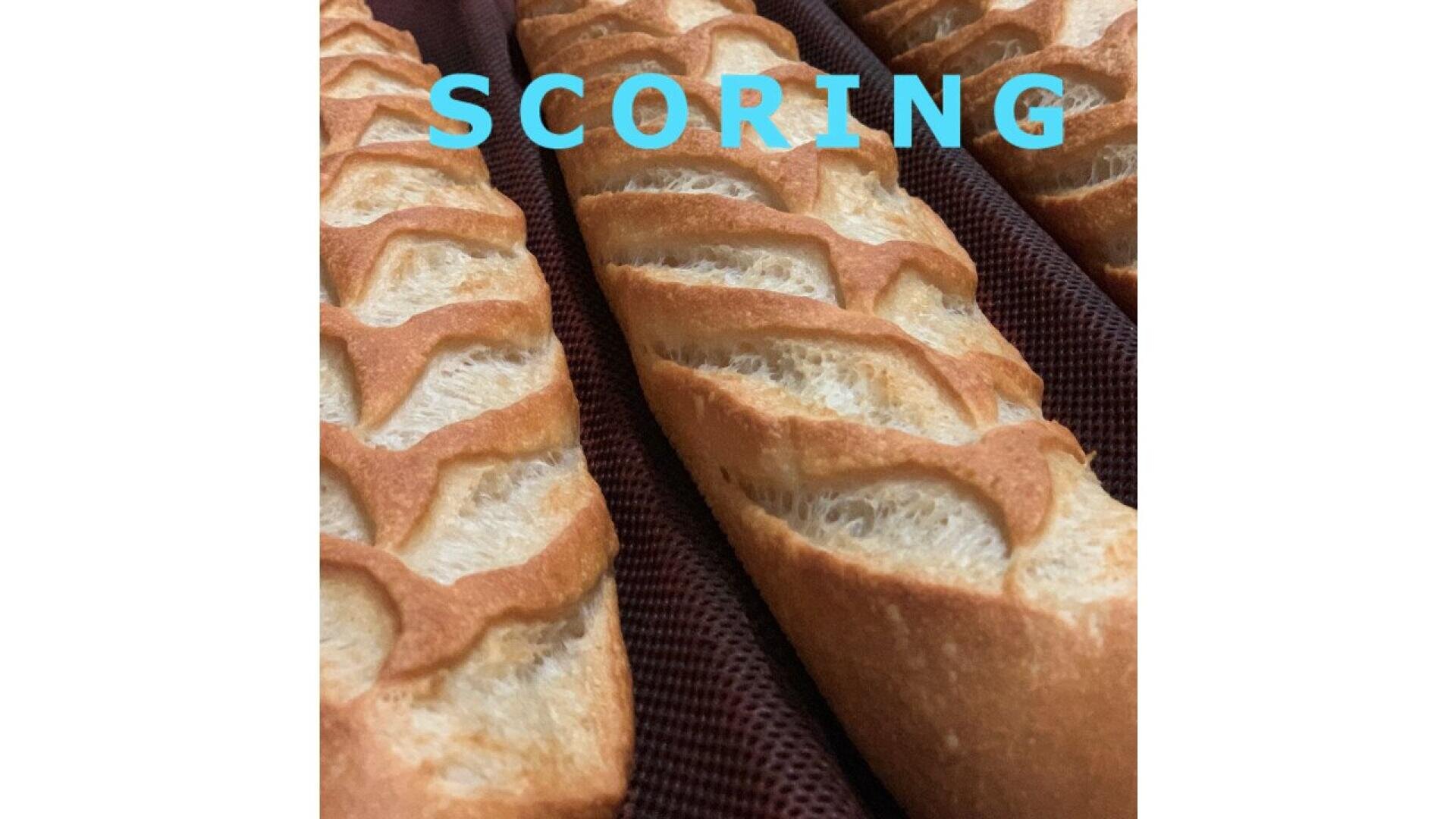 Drei Baguettes mit tiefen, diagonalen Schnitten auf der Oberseite liegen auf einer Backmatte. Das Wort SCORING erscheint in fetten blauen Buchstaben über den Broten.