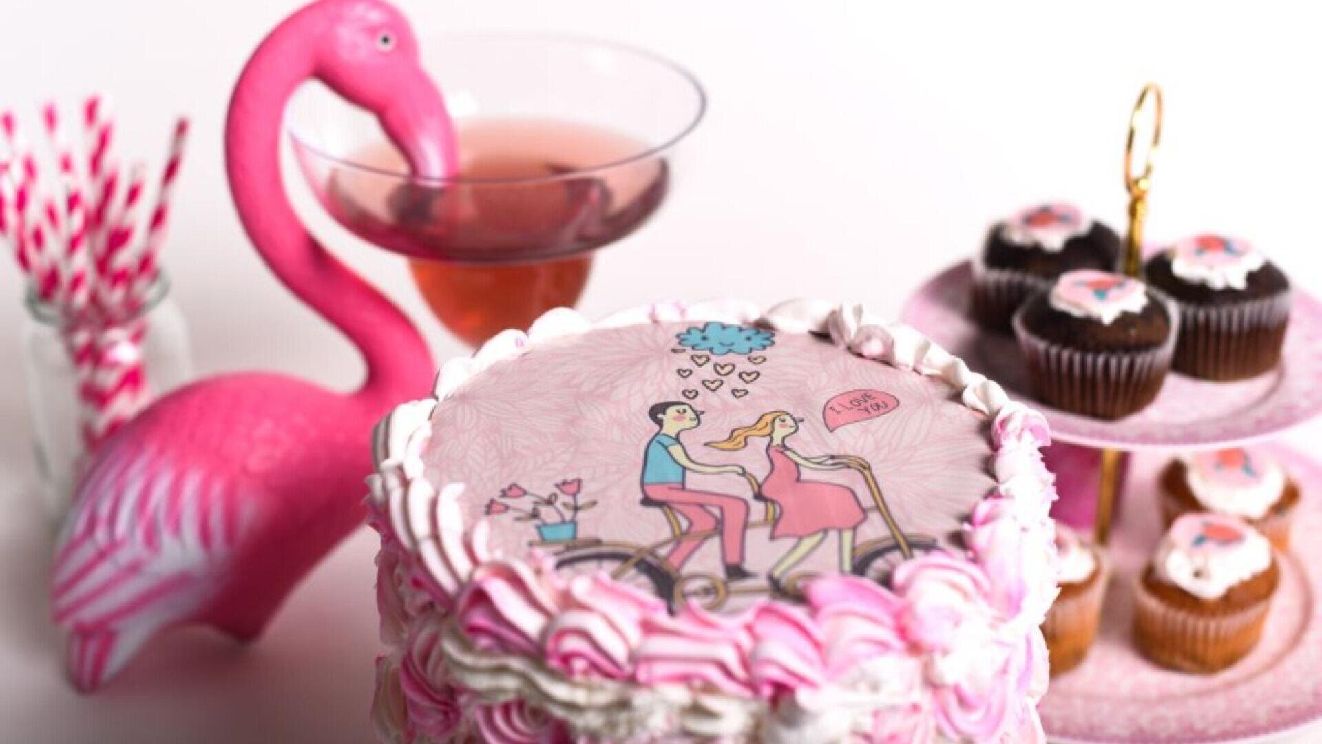 Ein rosafarbenes Dessert-Setup mit einer Torte, die mit einem Cartoon-Paar auf einem Fahrrad verziert ist, einer rosafarbenen Flamingo-Dekoration, einem Getränk in einem Cocktail-Glas, rosa gestreiften Strohhalmen und Cupcakes auf einem Ständer mit mehreren Ebenen.