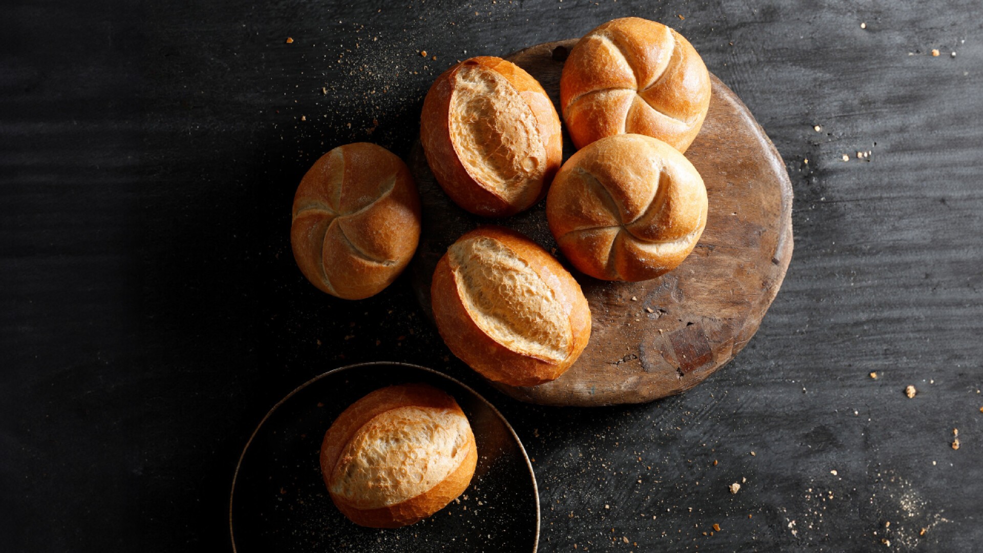 Six round bread rolls are arranged on a rustic wooden board and a dark surface. Some rolls have a split pattern while others have a star pattern. Bread crumbs are scattered around.