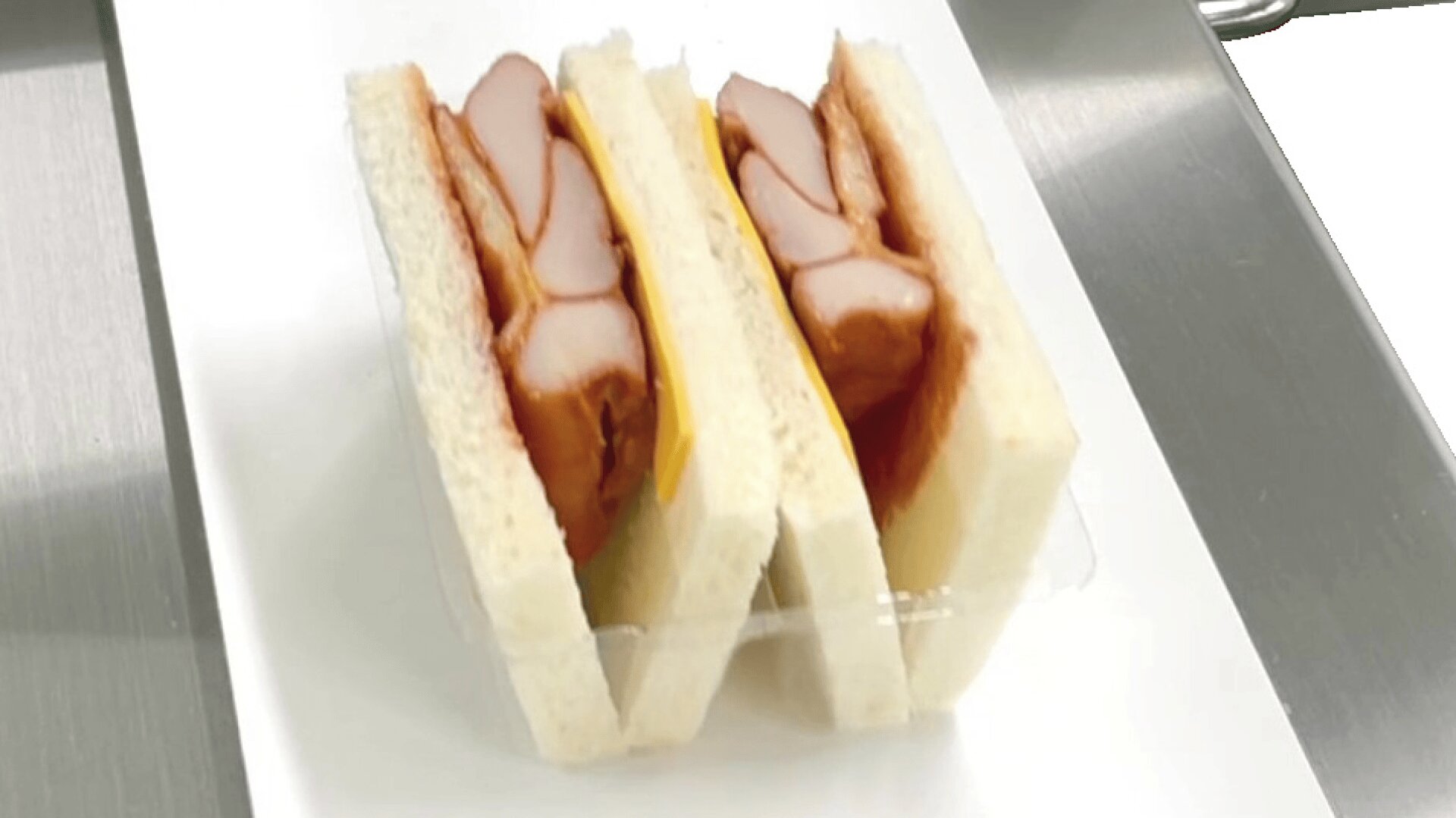 Zwei Sandwiches mit dicken Wurstscheiben und einer Scheibe Käse zwischen Weißbrot, serviert auf einem weißen, rechteckigen Teller.