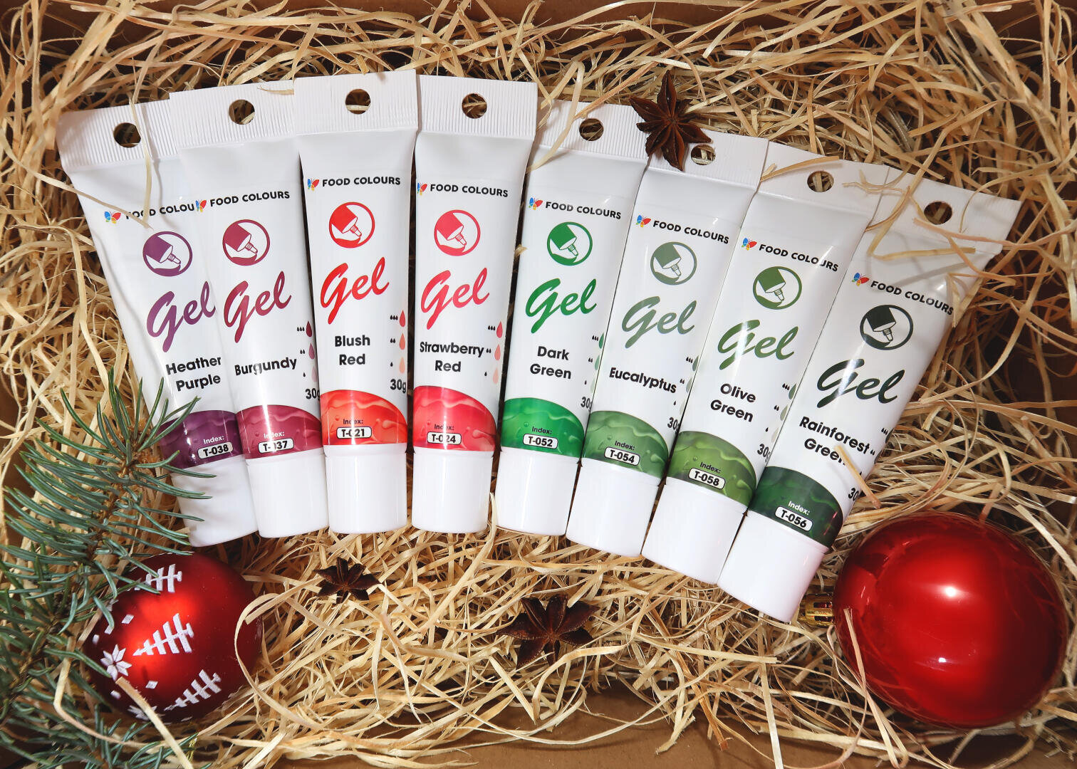 Seven tubes of gel food colors in various shades are displayed on straw with festive decorations, including pine branches and red ornaments. The colors range from purple and red to different shades of green.
