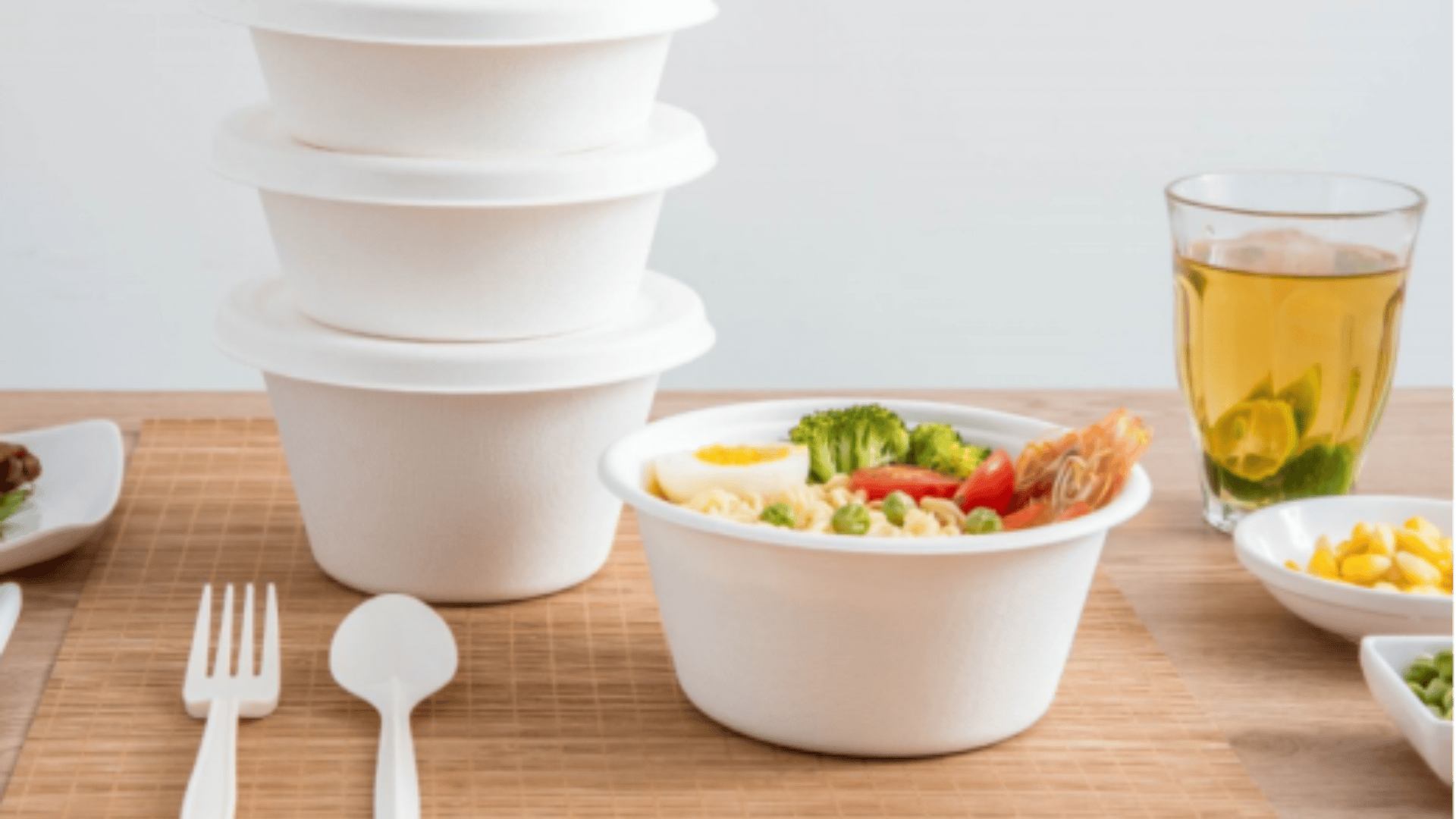 A table with white disposable bowls, one filled with noodles, shrimp, broccoli, and vegetables. Three stacked bowls, a glass of green tea, a fork, and a spoon are also on the table.