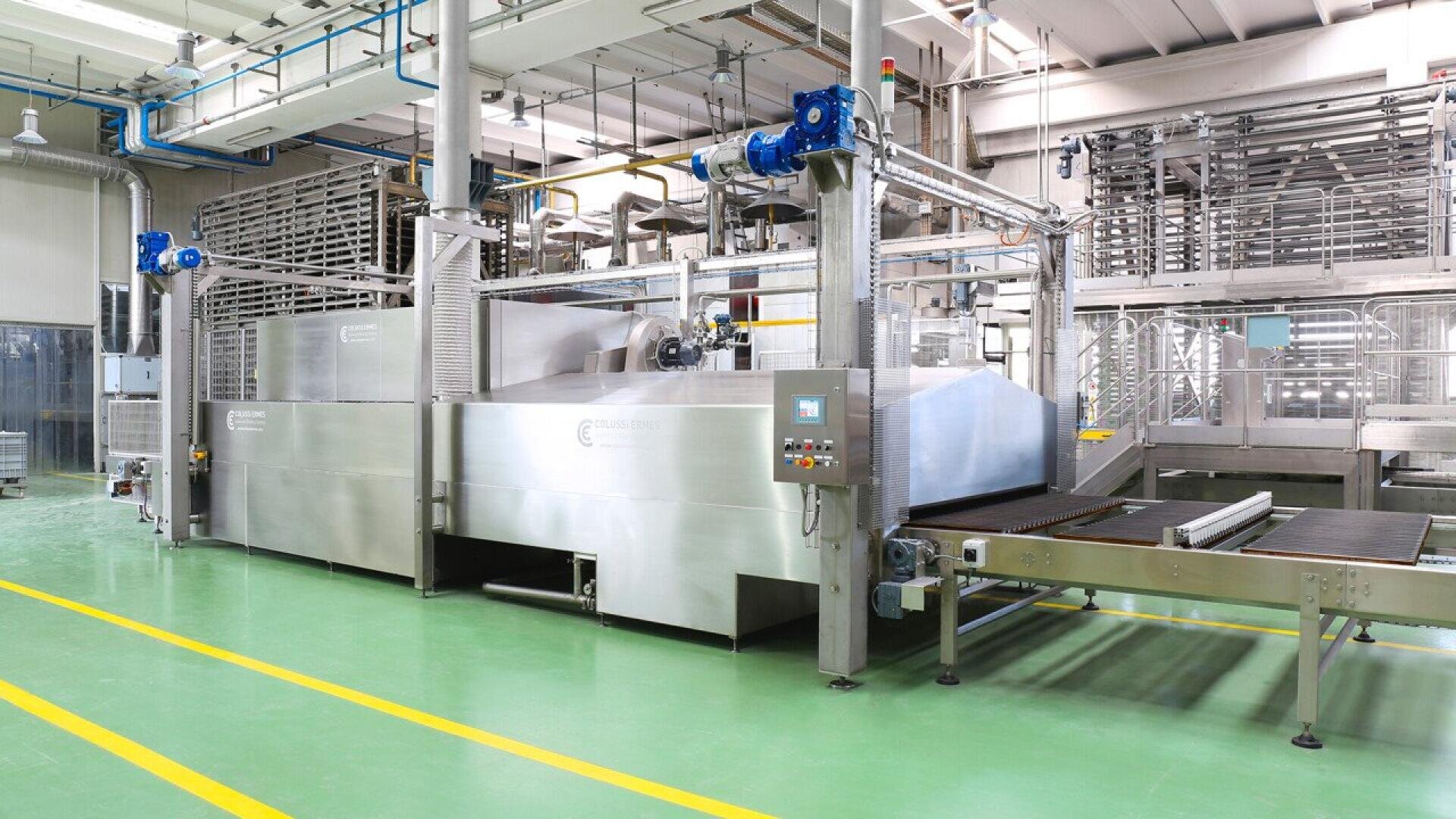 A large industrial food processing machine with stainless steel surfaces and conveyor belts is set in a clean, modern factory with green floors and overhead pipes.