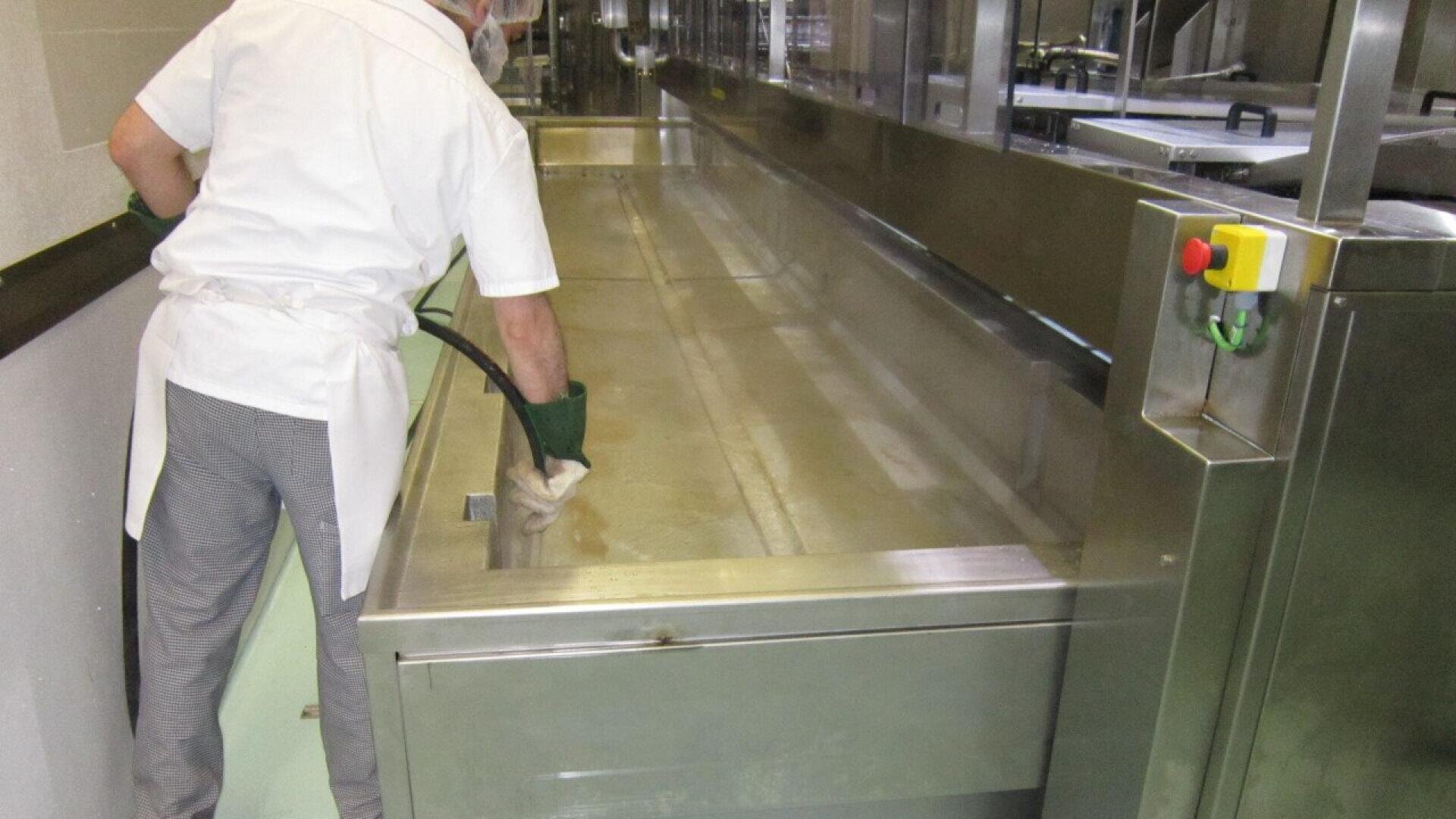 Eine Person, die Schutzhandschuhe und eine weiße Uniform trägt, reinigt oder spült einen großen Edelstahltrog in einer gewerblichen oder industriellen Küche. Die Person hält einen Schlauch in der Hand und schaut von der Kamera weg.