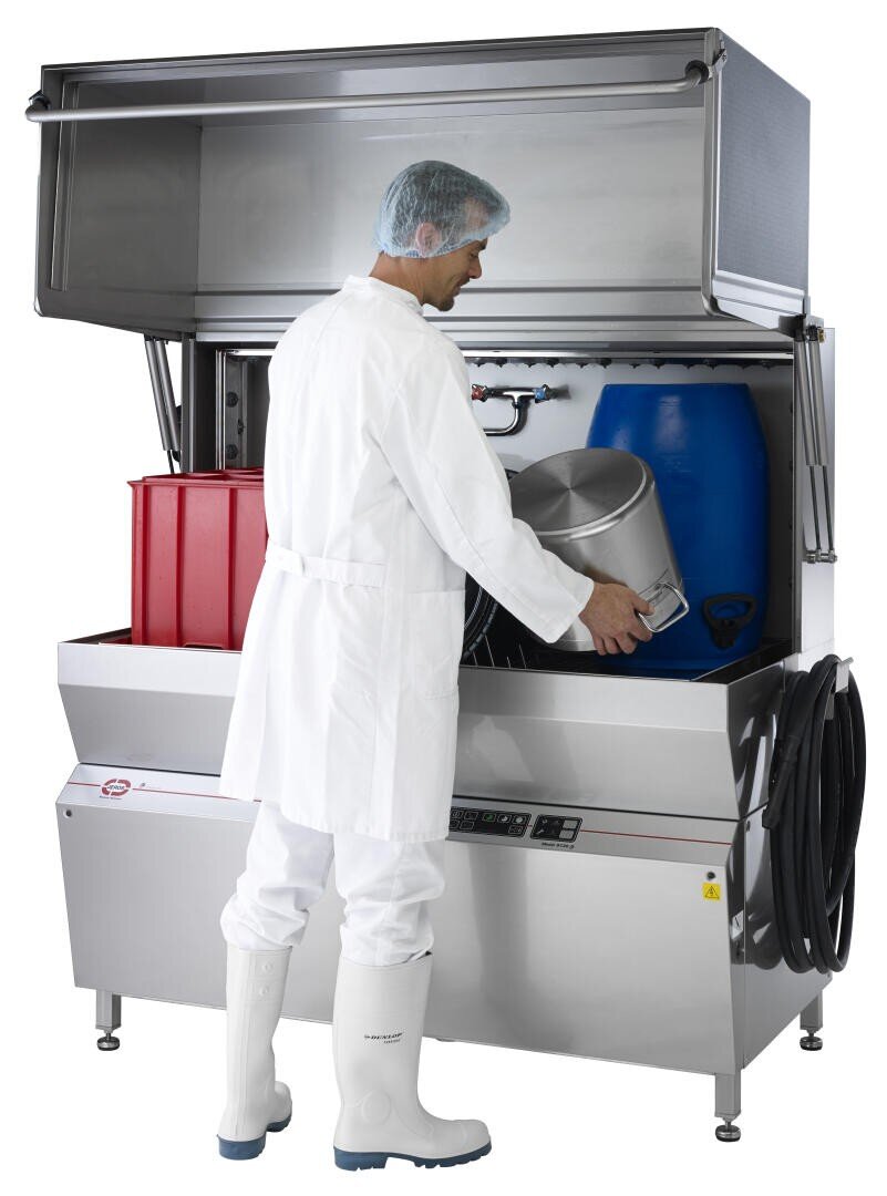 Eine Person in weißer Schutzkleidung und Stiefeln stellt einen großen Metallbehälter in eine industrielle Waschmaschine mit roten und blauen Kunststoffbehältern im Inneren. Der Deckel der Maschine ist geöffnet, und an der Seite sind verschiedene Schläuche zu sehen.