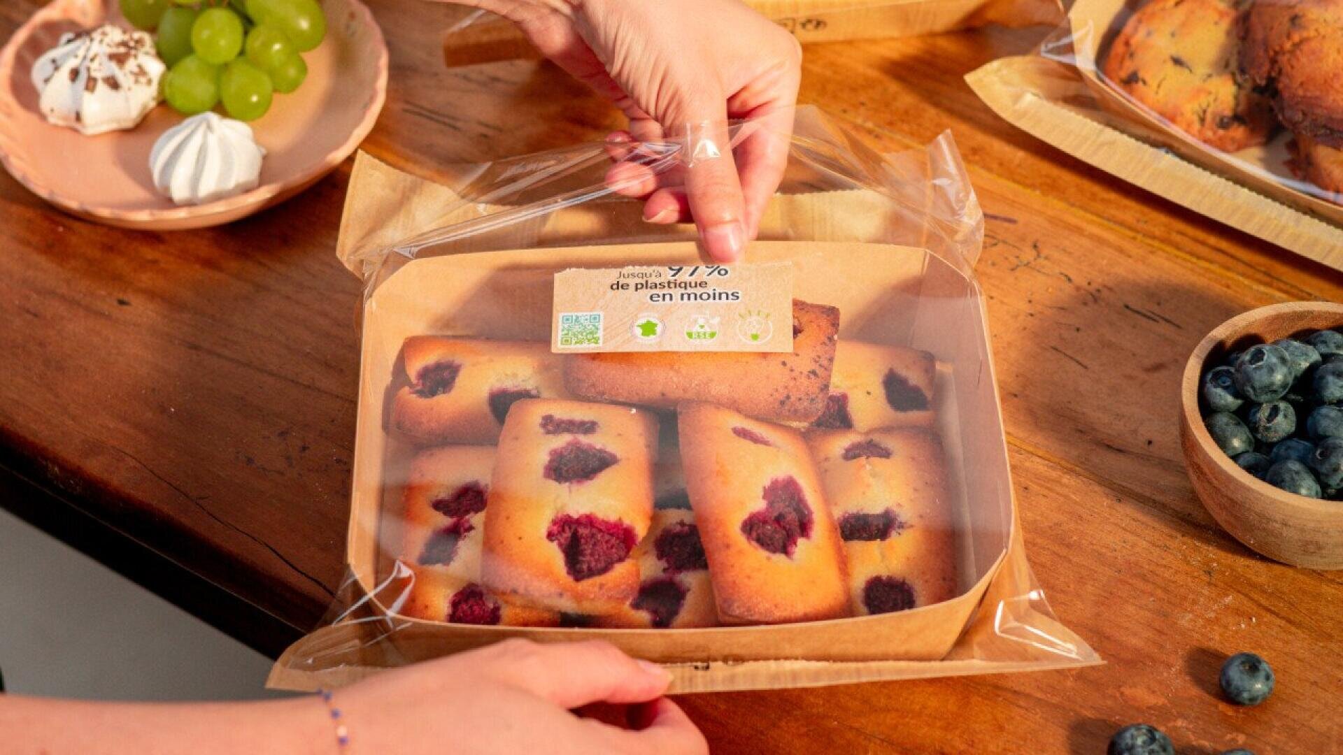 Eine Person öffnet einen durchsichtigen Plastikbehälter mit rechteckigen Backwaren mit Himbeeren, der auf einem Holztisch mit Weintrauben, Keksen und Blaubeeren in der Nähe steht.